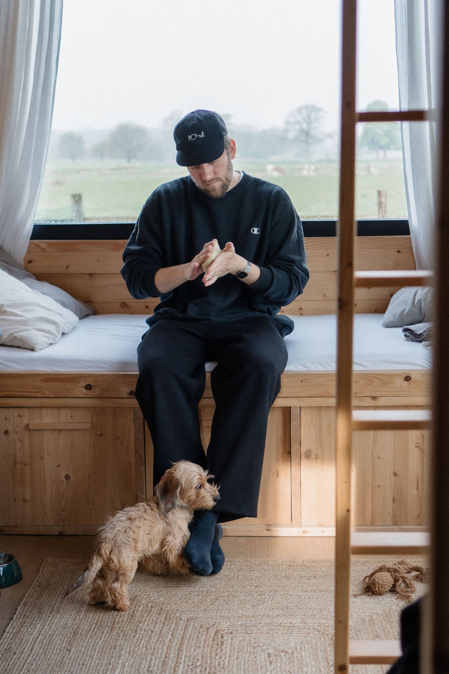 Man in black Champion sweatshirt sits on wooden bed with small fluffy dog in cozy cabin interior.