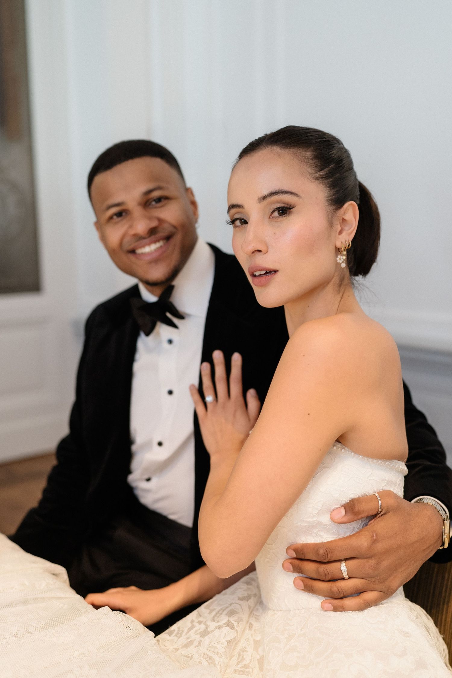 Smiling groom in black tuxedo embracing bride in strapless lace wedding dress while seated on floor.