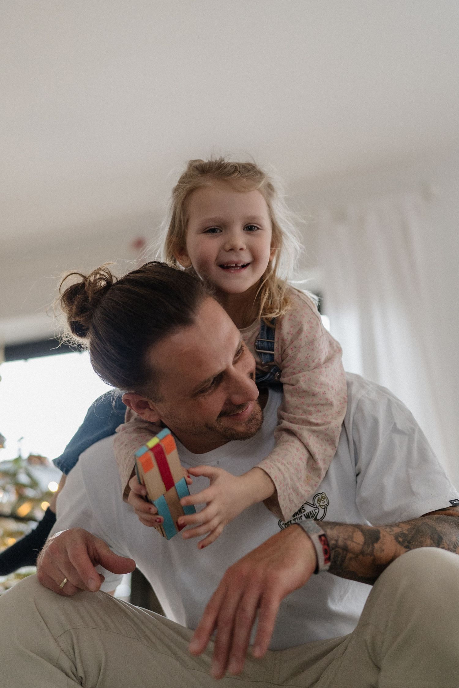 A tattooed father plays with his smiling blonde toddler daughter, who climbs on his back holding colorful toy blocks.