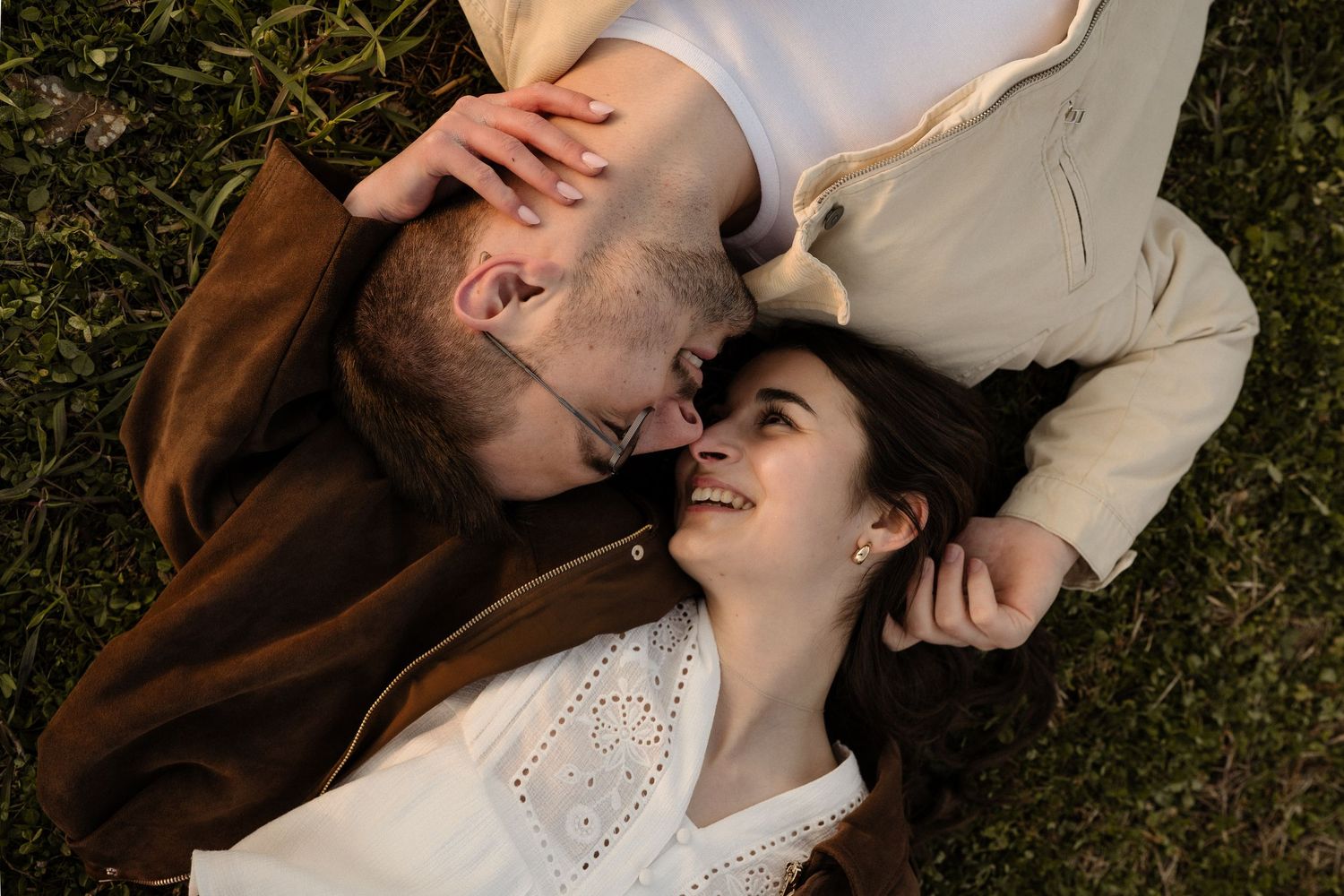 Romantic couple lying on grass, sharing a tender kiss moment, woman smiling joyfully in outdoor setting.