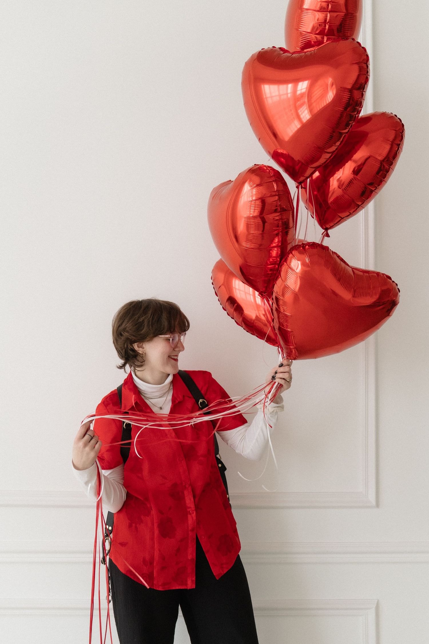 Smiling woman in red vest looking at red heart-shaped balloons she holds up.