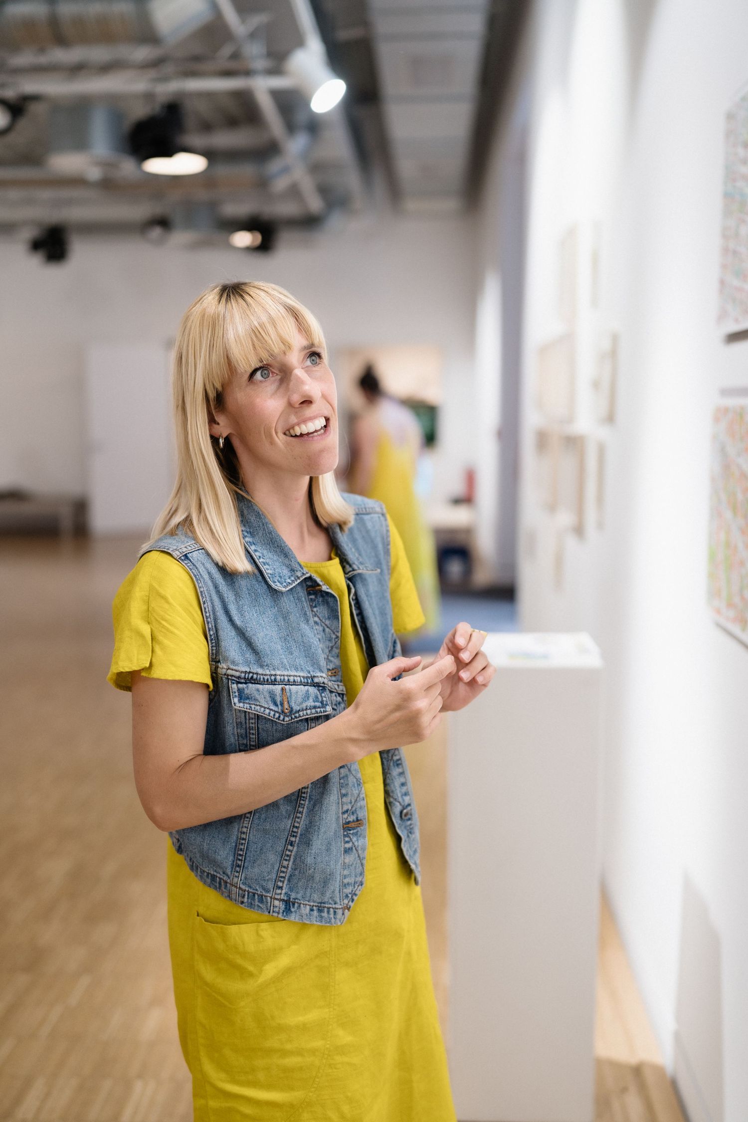 Smiling blonde woman in yellow dress and denim vest admiring artwork on white gallery walls.