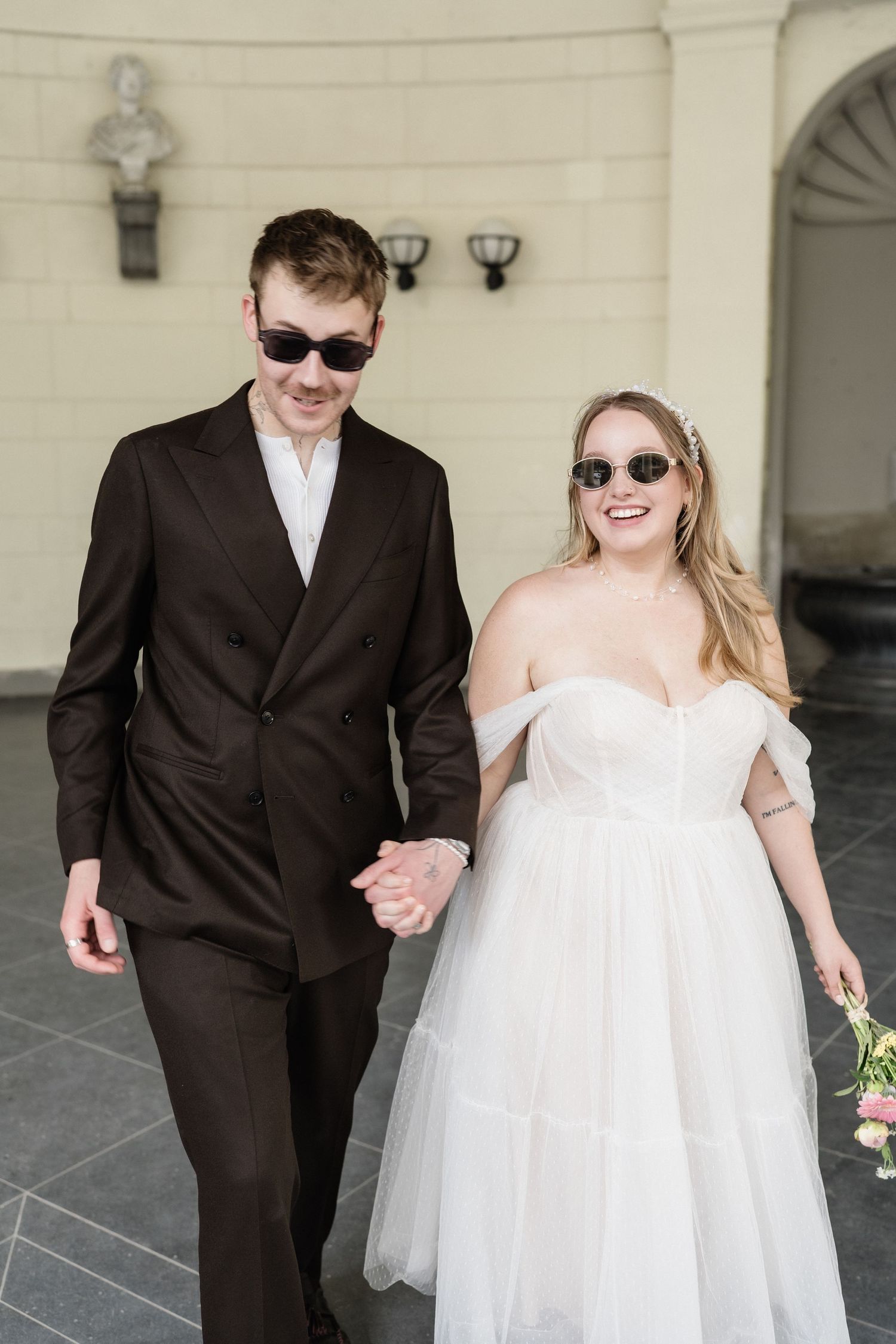 Smiling bride in off-shoulder tulle gown and groom in dark suit walk hand in hand wearing sunglasses.