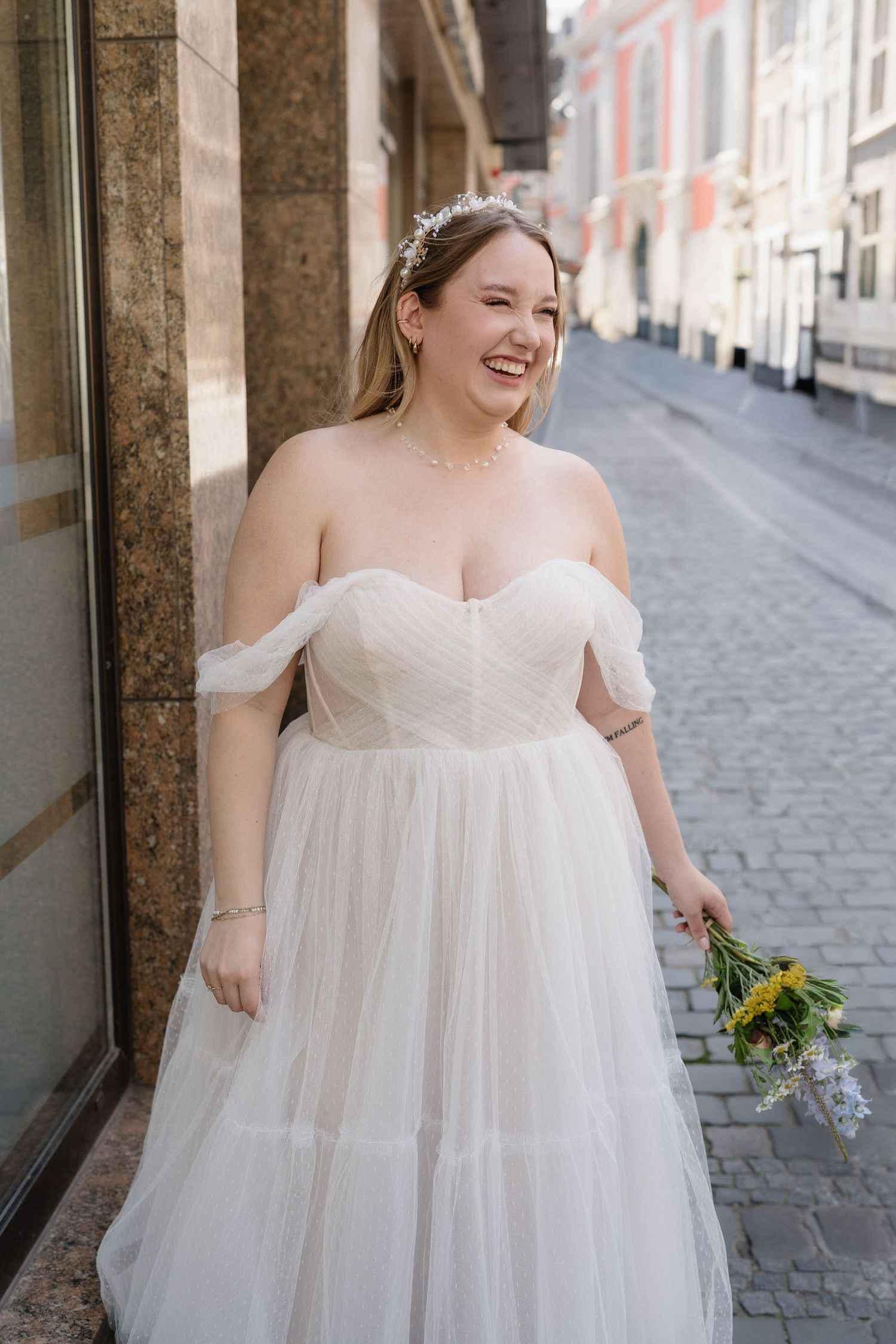 Smiling bride in off-shoulder tulle wedding gown holding sunflower bouquet on a cobblestone street.