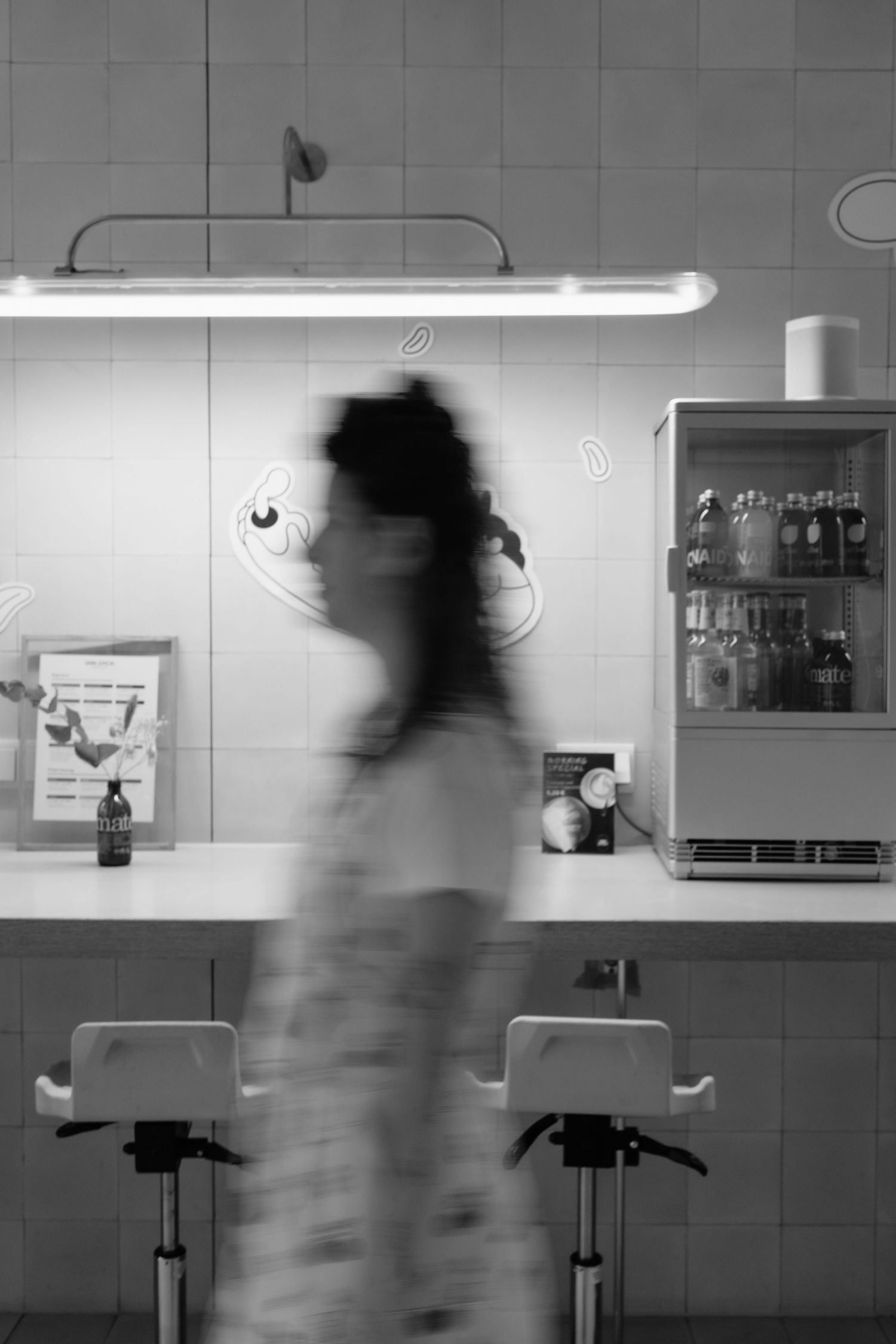 Blurred woman walking past a cafe counter with neon lights and refrigerator.