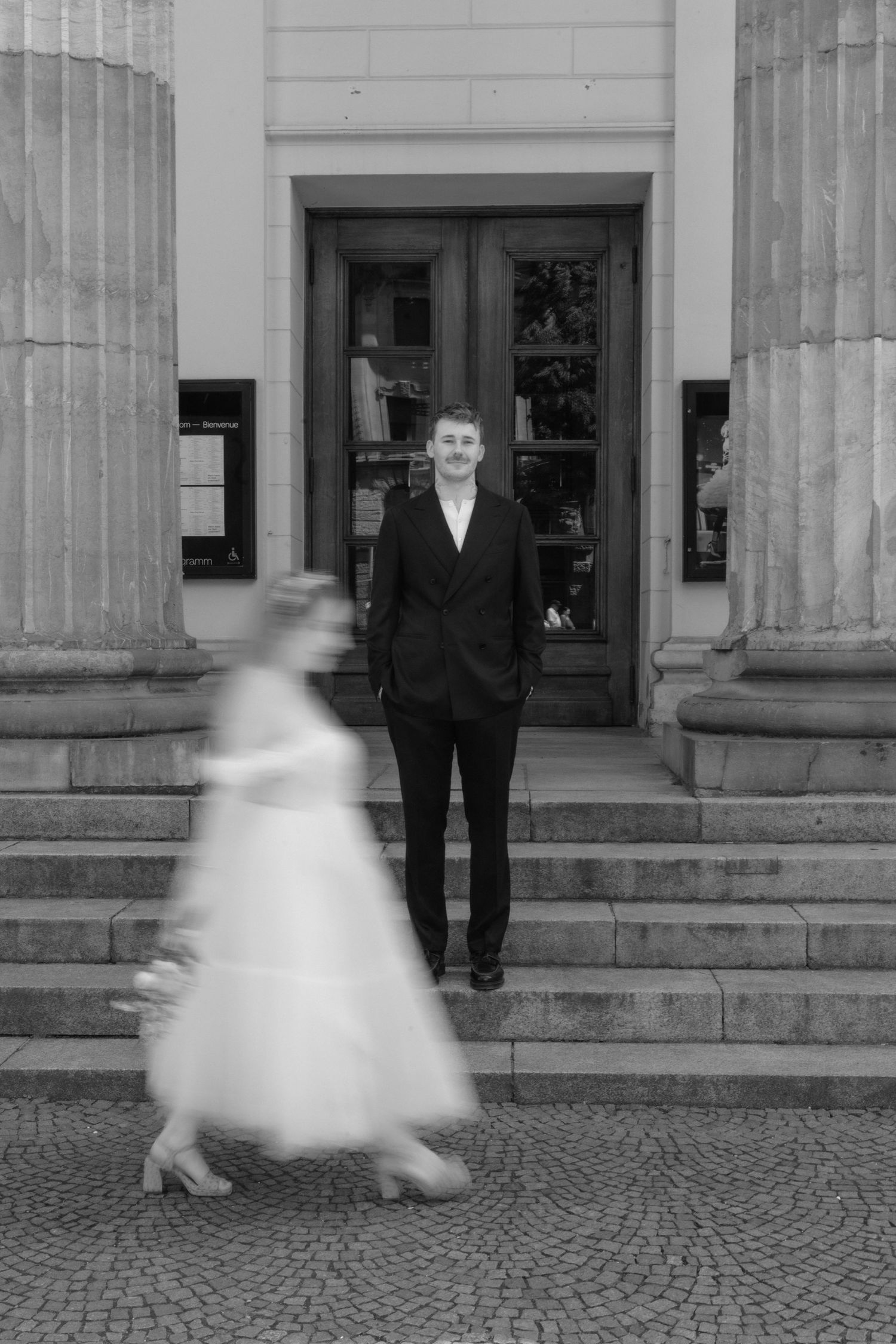 Black and white photo of groom standing on steps as blurred bride in tulle dress walks past.