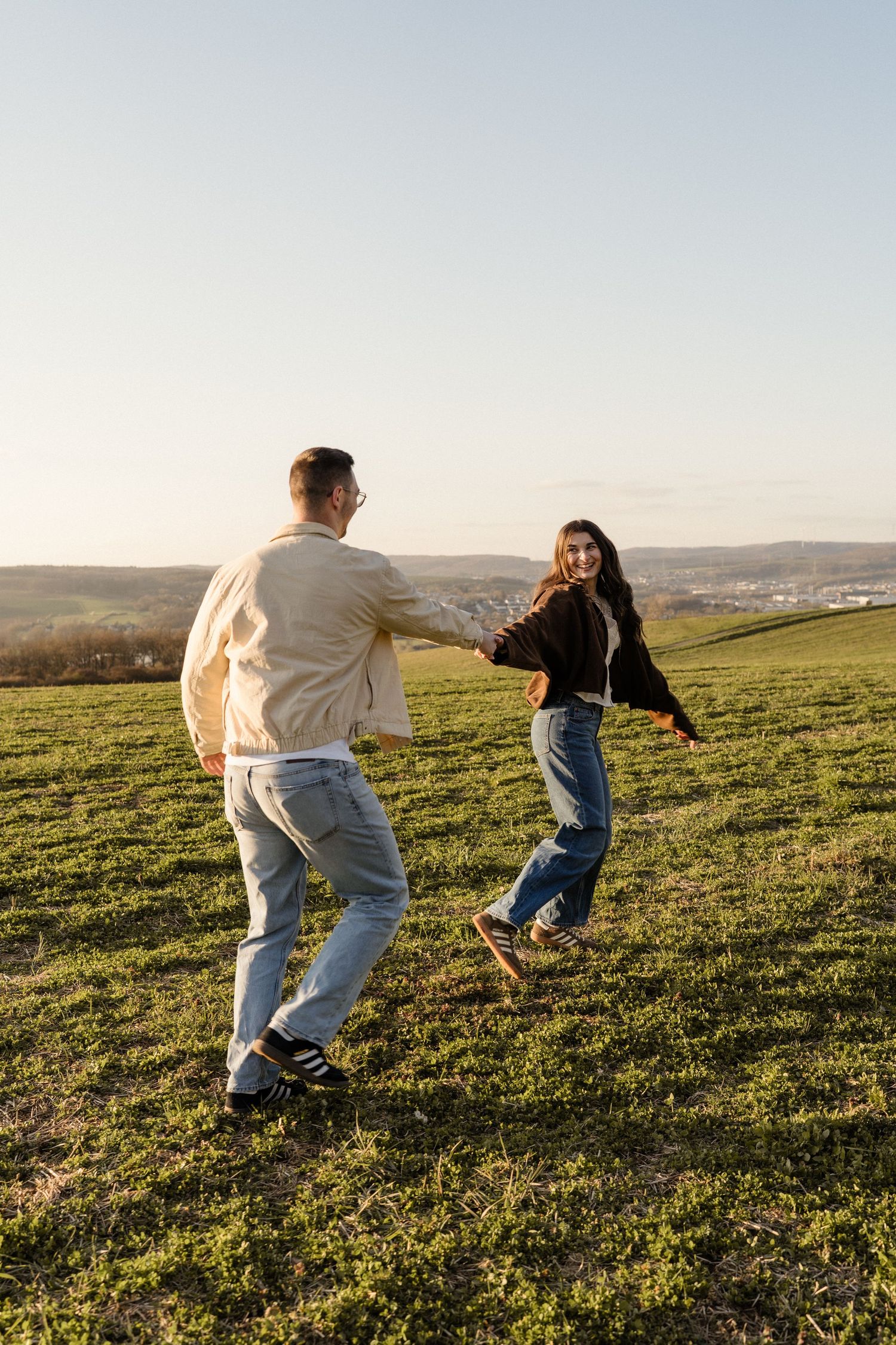 Happy couple holding hands and dancing playfully on a green hillside at sunset.