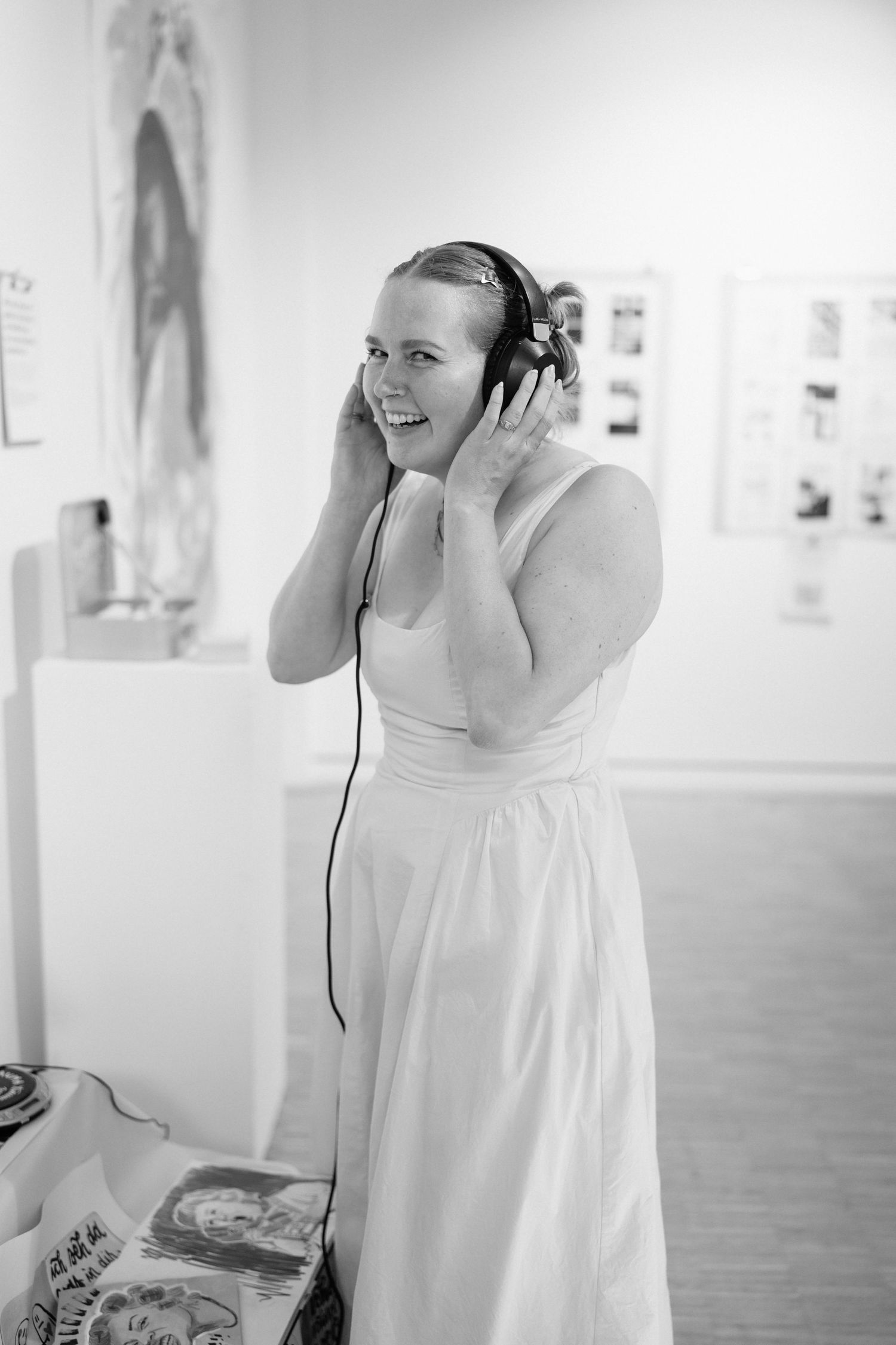 Woman in white dress smiling while wearing headphones in an art gallery, black and white photo.