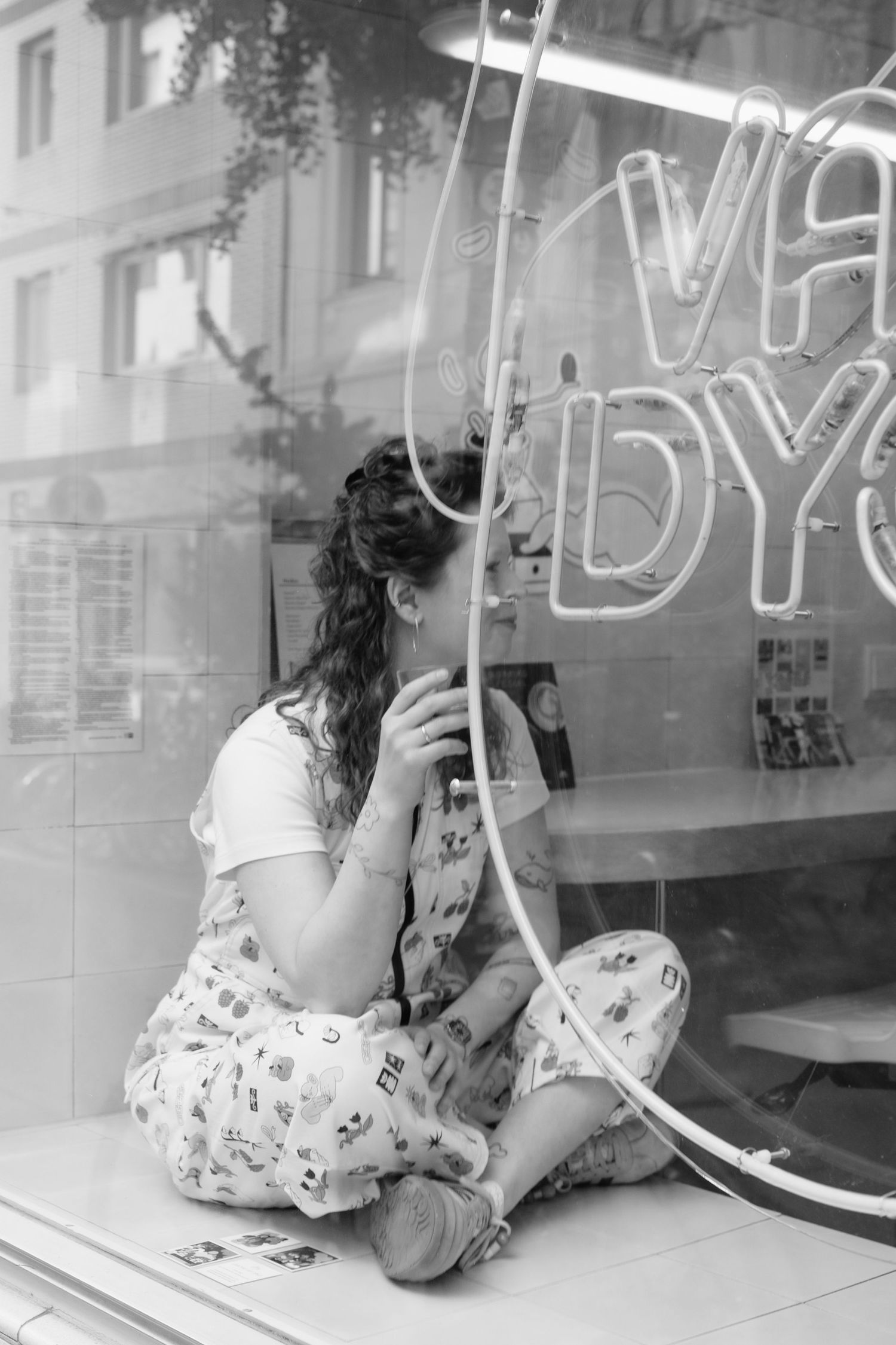 Young woman sitting cross-legged in cafe window with neon sign reflection.