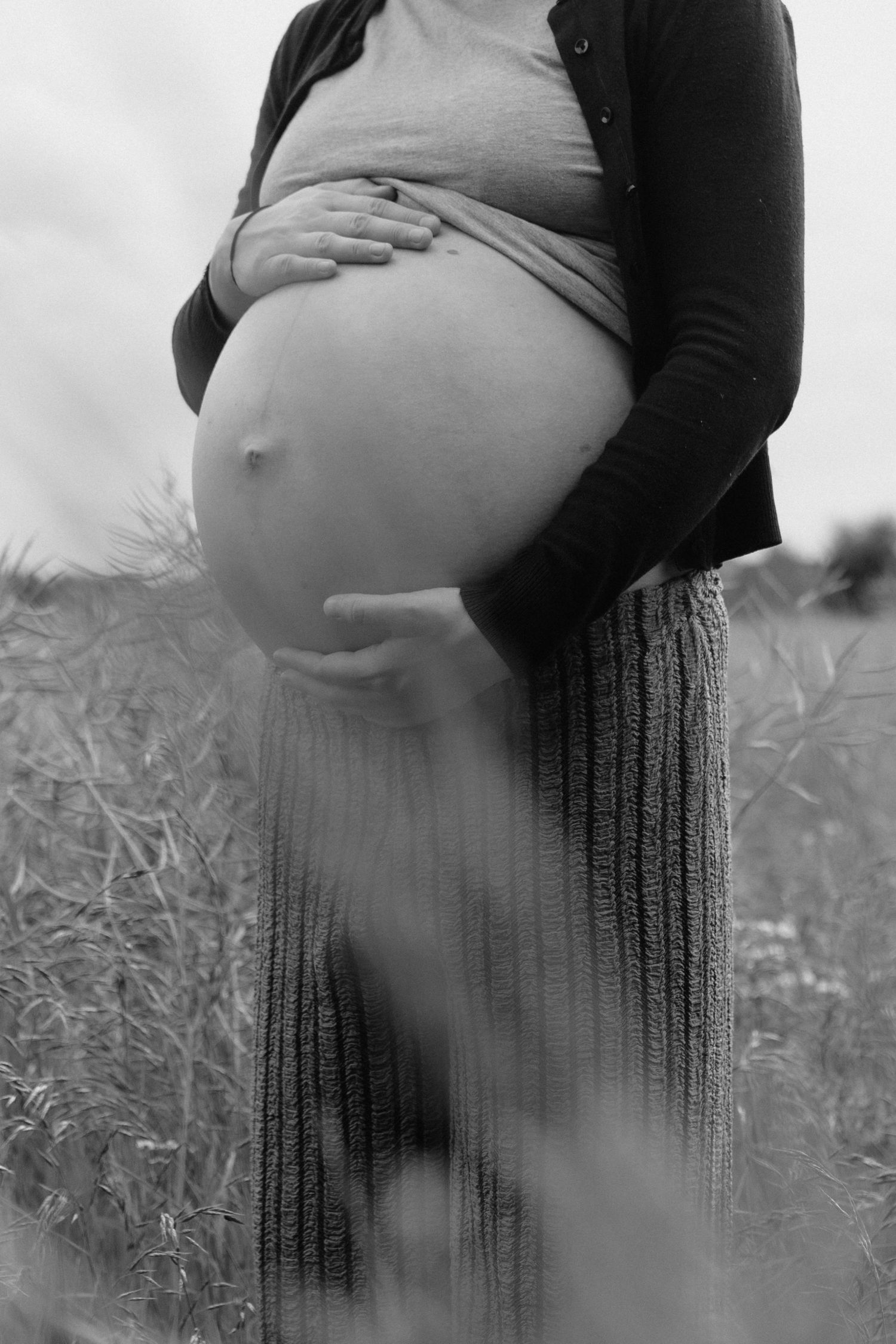 Black and white photo of pregnant woman cradling bare baby bump while standing in a field.