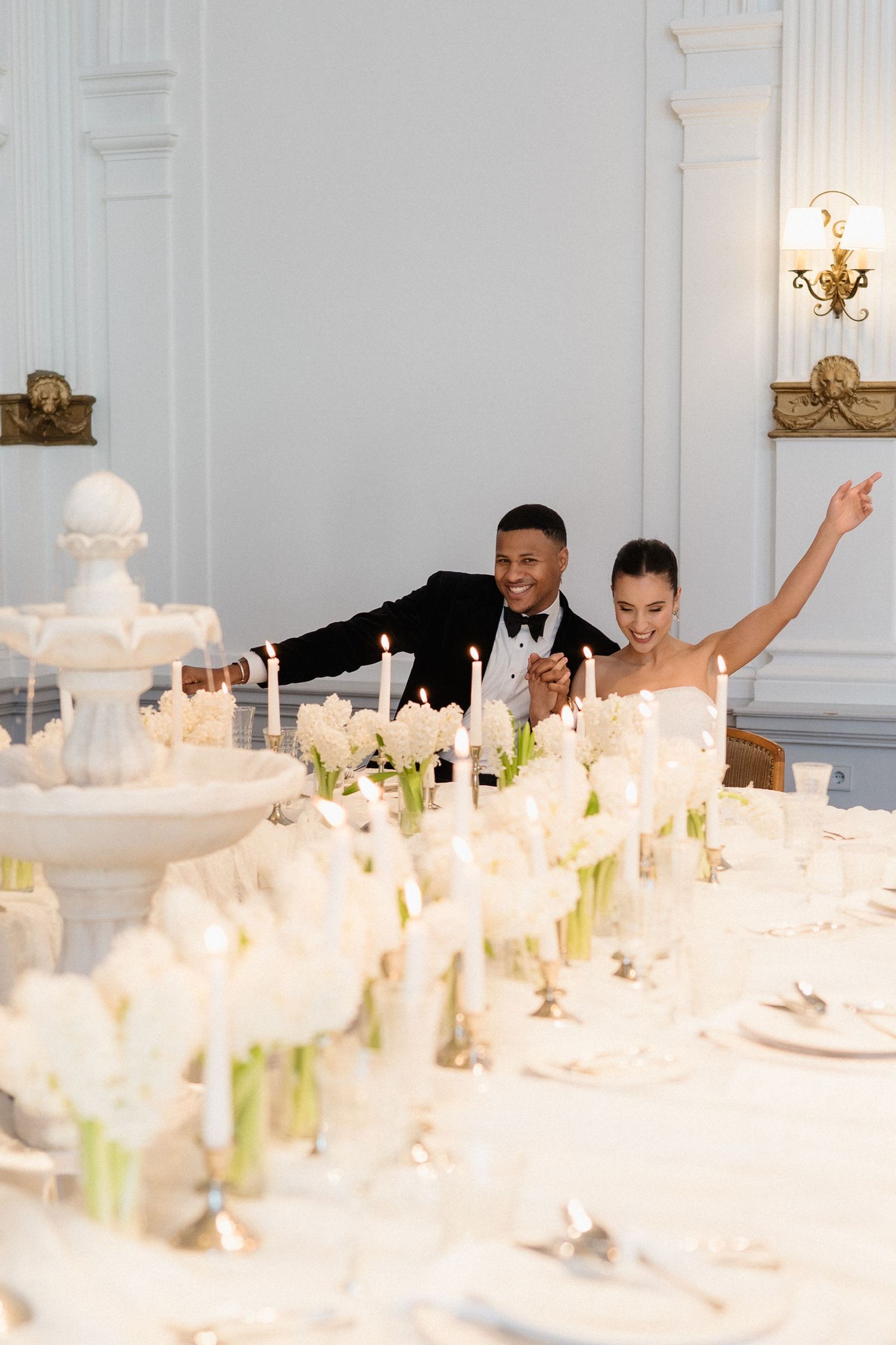 Joyful interracial couple celebrating at elegantly decorated wedding reception table with white florals and candles.