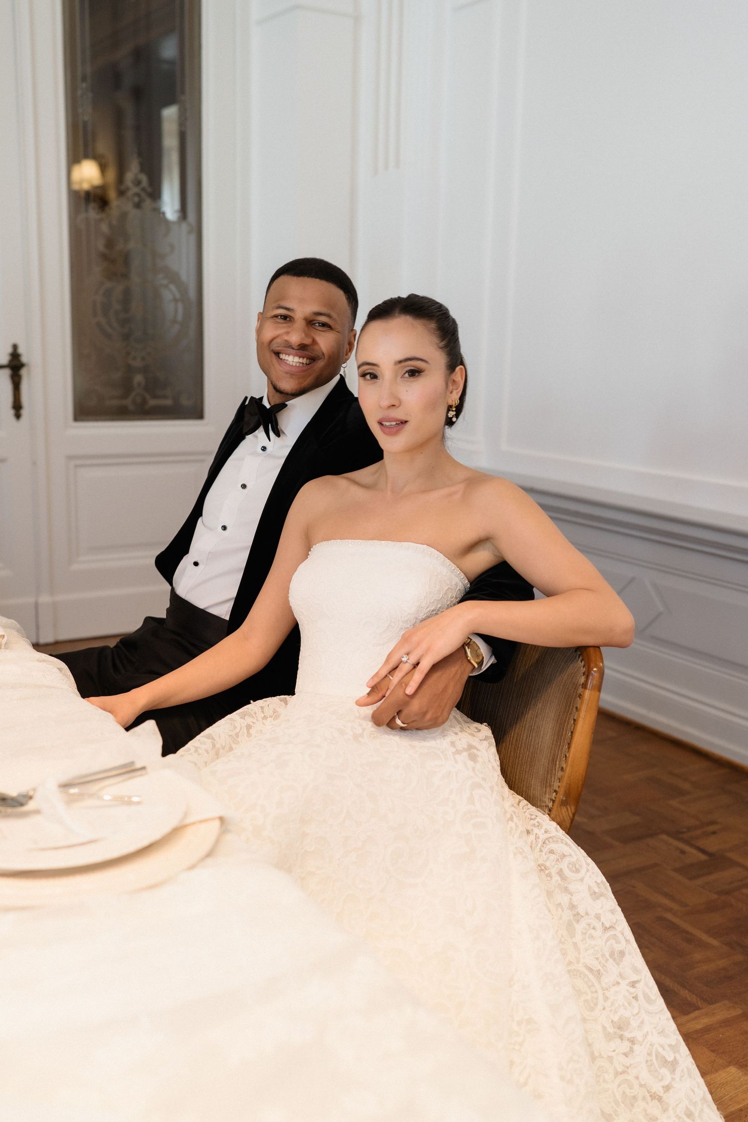 Elegant interracial newlywed couple posing at wedding reception table in a classic white ornate room.