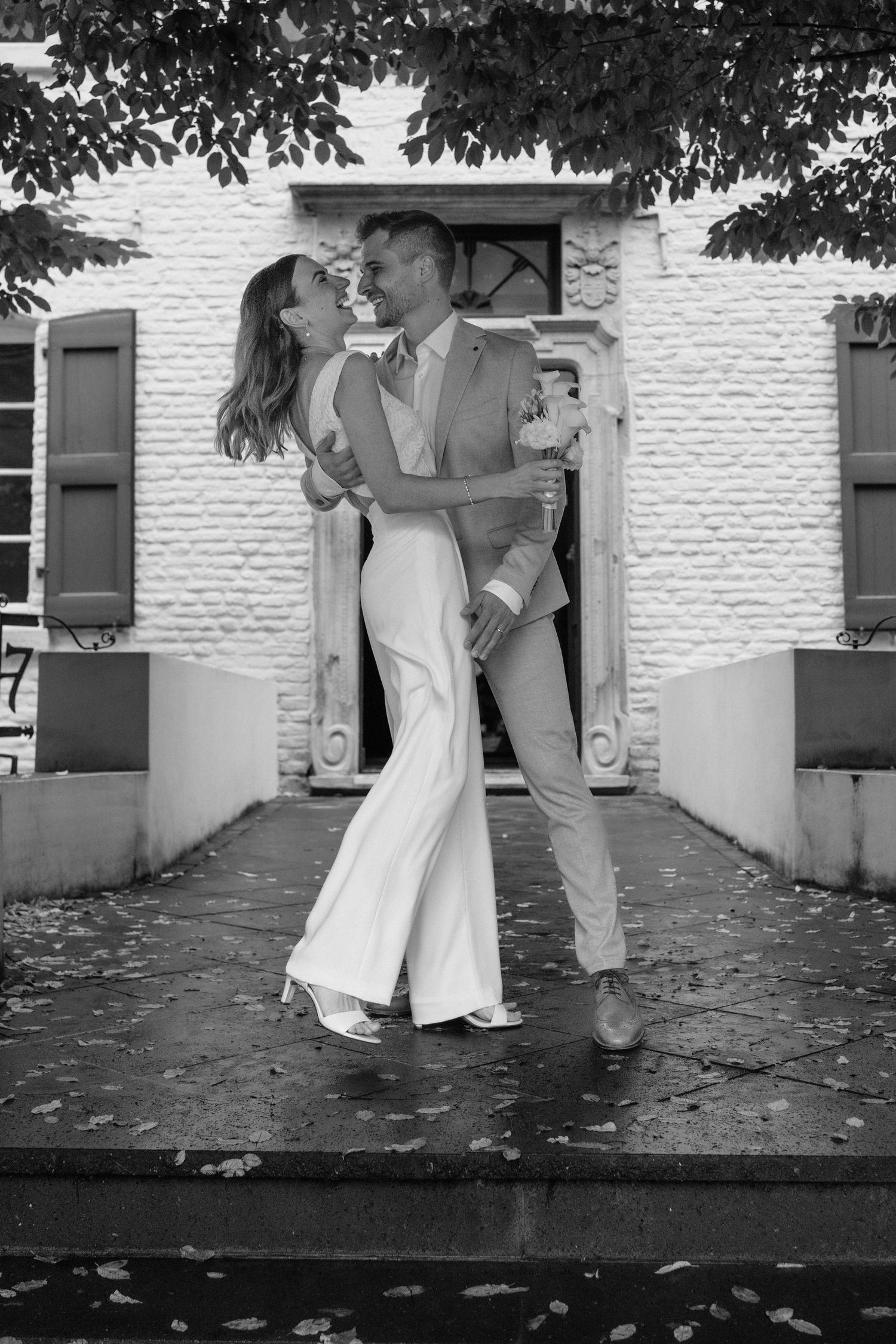 Black and white photo of a bride and groom embracing and laughing outside a white brick building.