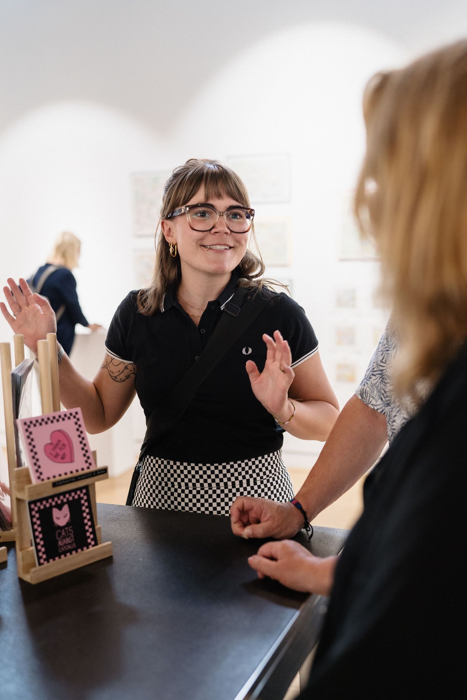 A smiling woman with glasses and tattoos gestures with both hands while talking to someone at a counter in an art gallery.
