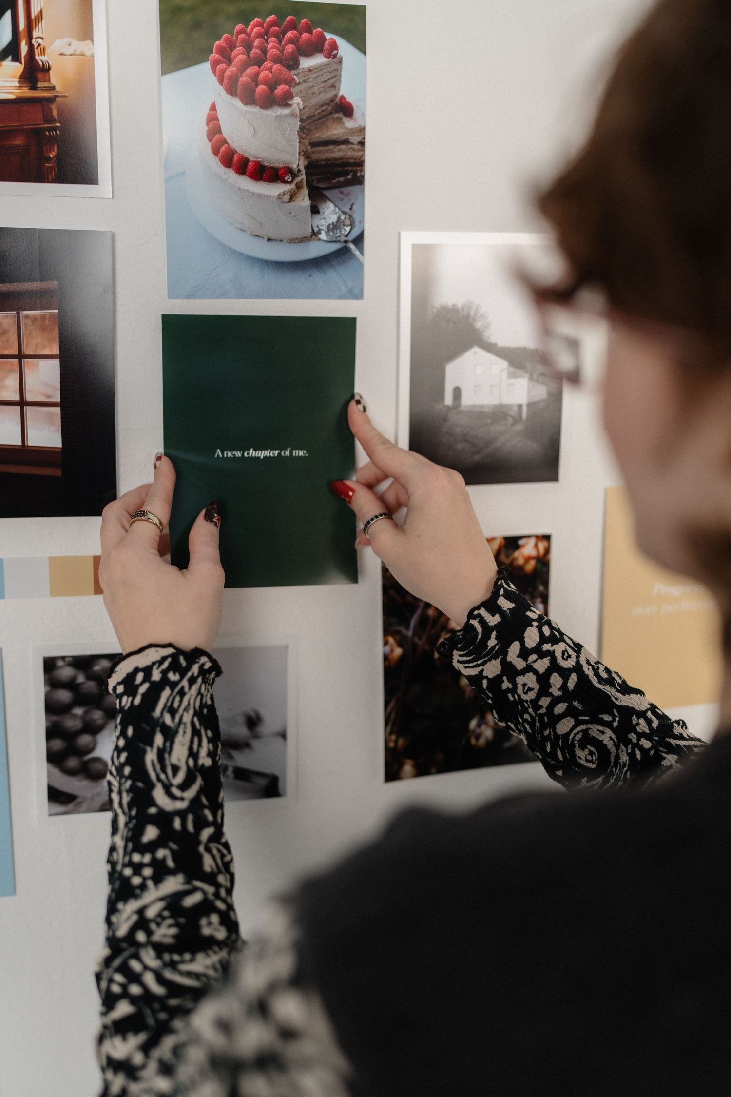 Woman arranging photos on a mood board wall featuring a raspberry cake image and green card reading 'A new chapter of me'.