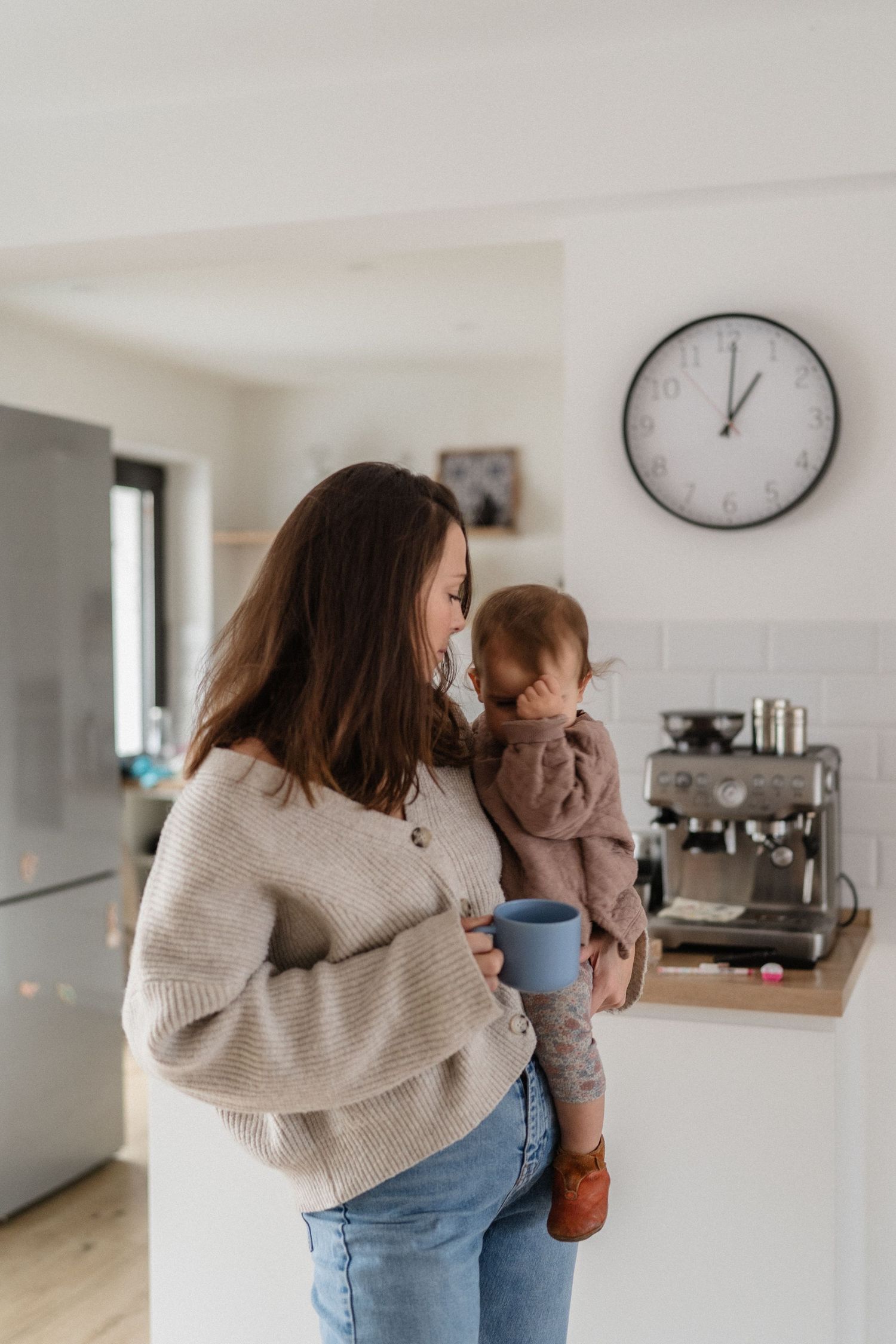 A mother holds her toddler while drinking coffee in a modern white kitchen with a wall clock in the background.
