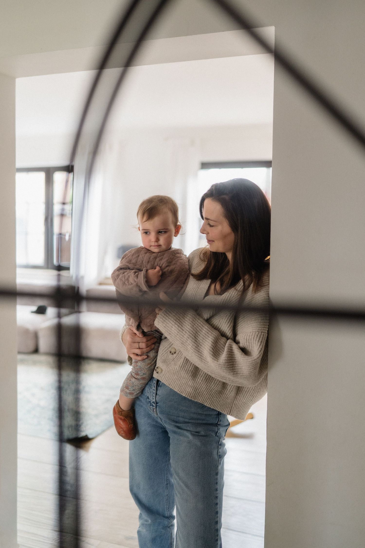 A young mother in a beige sweater and jeans holds her baby in a bright, modern living room.