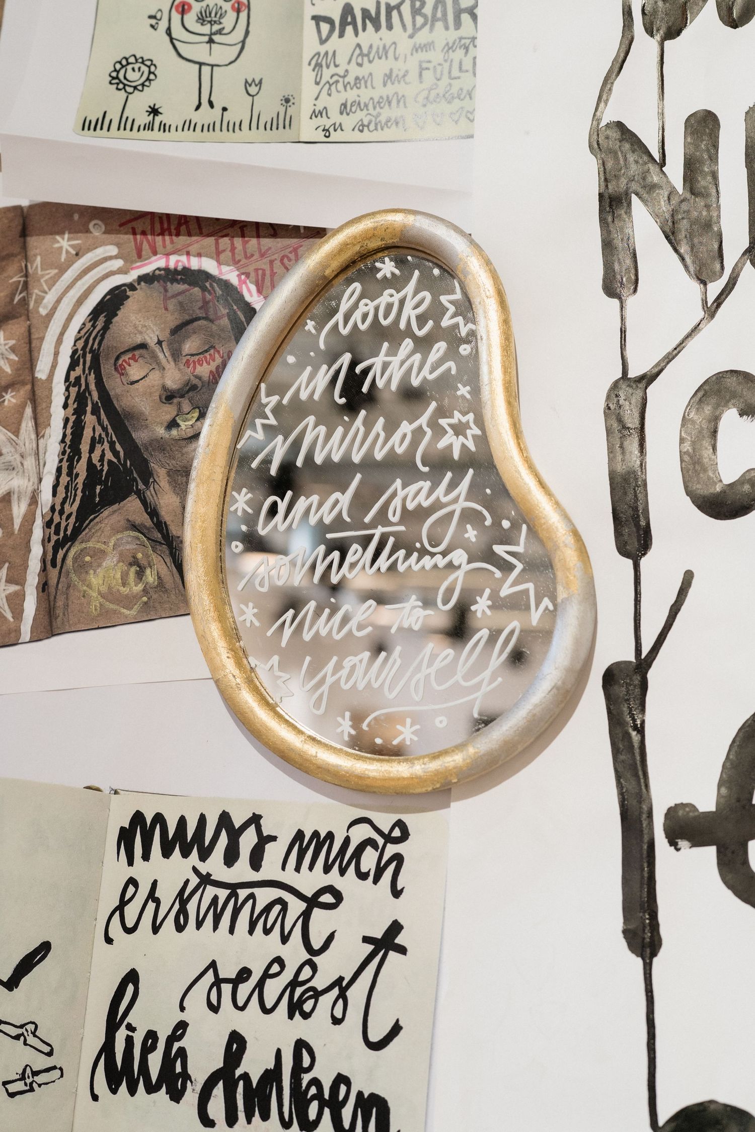 Gold pear-shaped mirror with motivational calligraphy text on a wall collage with artwork and German typography prints.