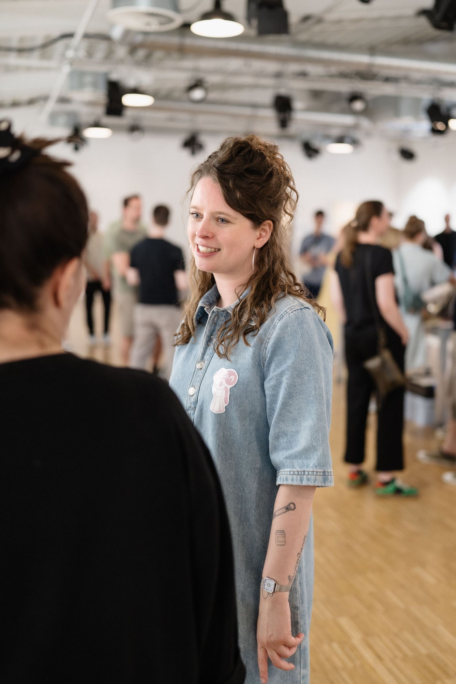 Smiling woman in denim dress with tattoos at a gallery event, surrounded by people in a bright modern space.