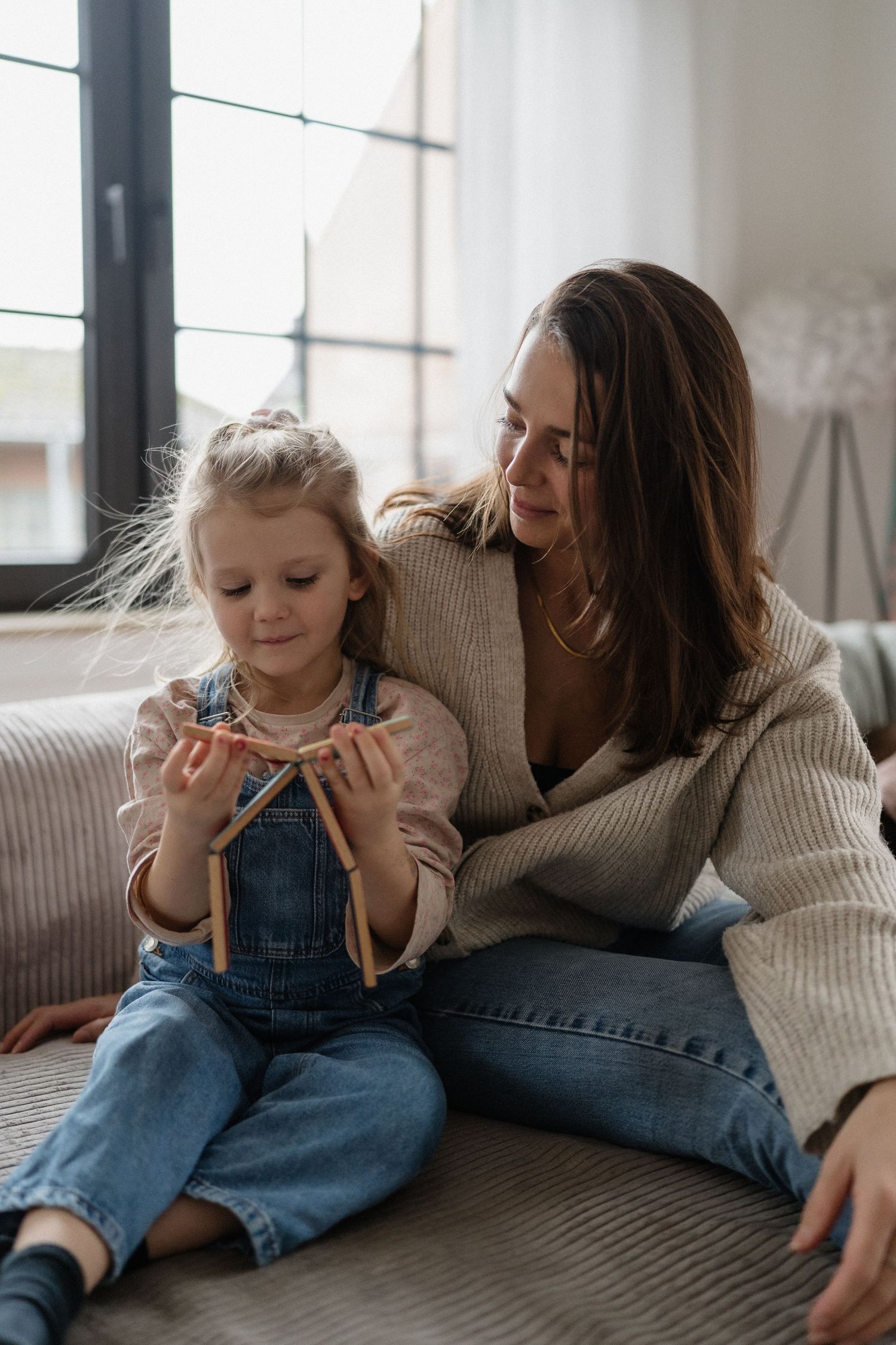 A mother and young daughter sit together on a couch, sharing a warm moment while the child plays with a ribbon bow.
