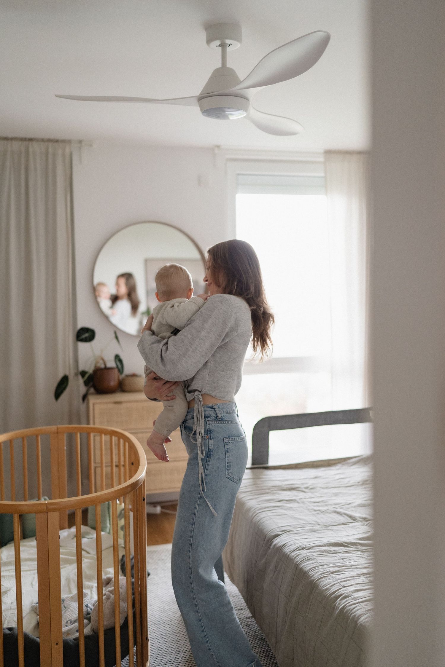 Young mother in jeans holding infant baby near wooden crib in bright modern bedroom.