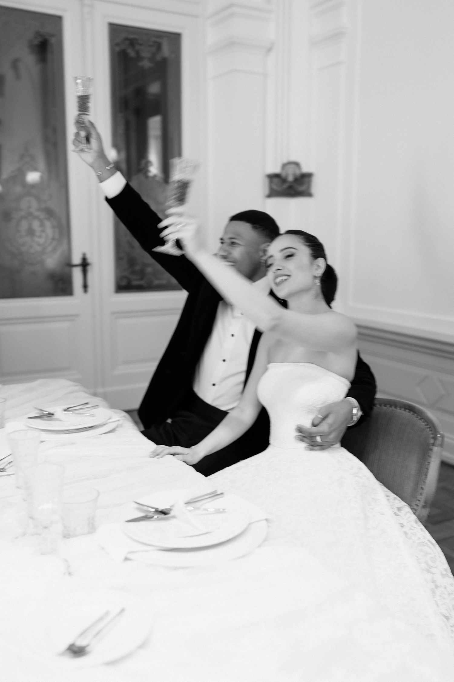 Black and white photo of bride and groom raising champagne glasses at wedding reception table.