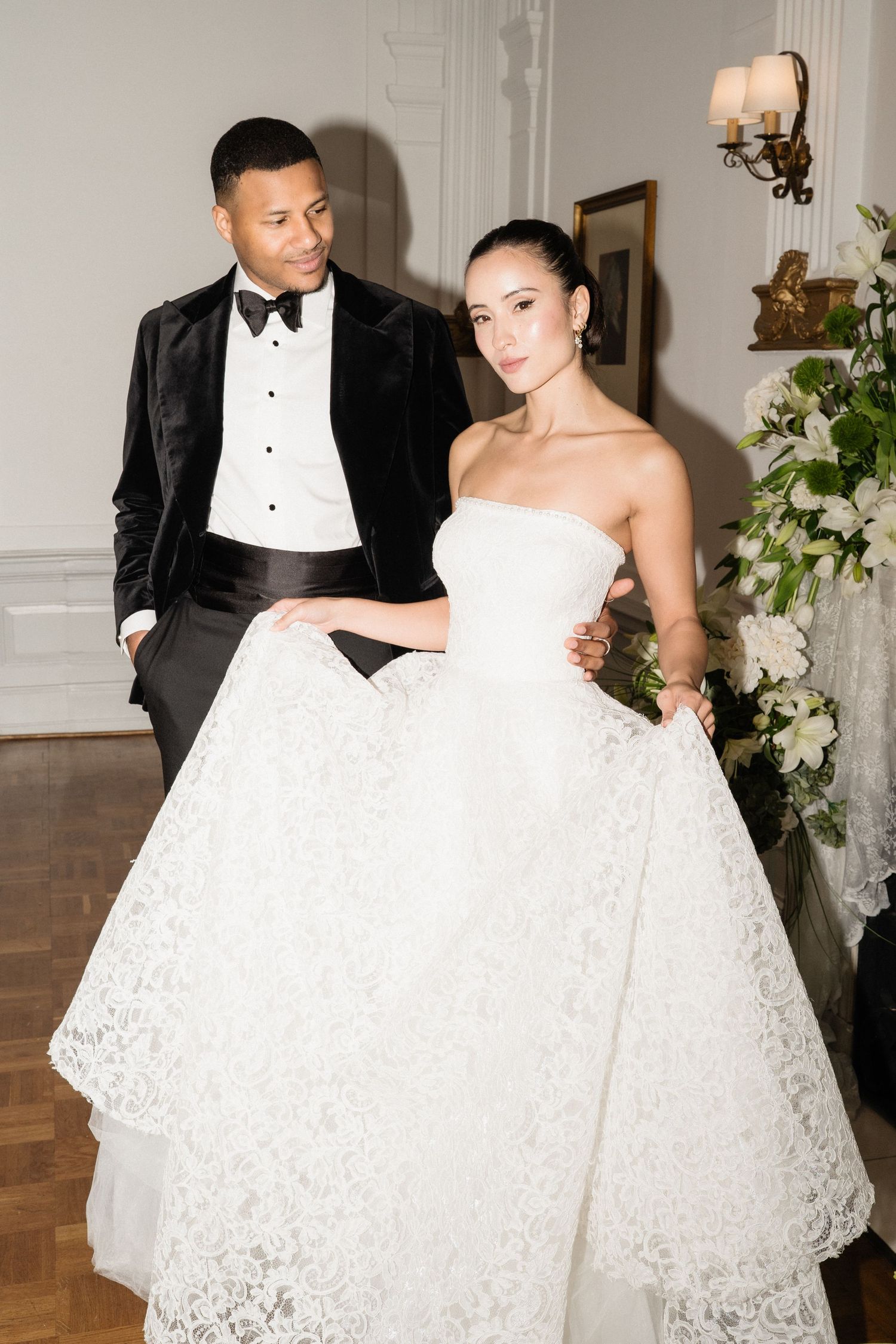 Elegant interracial couple posing in luxurious ballroom, groom in velvet tuxedo and bride in lace ball gown.