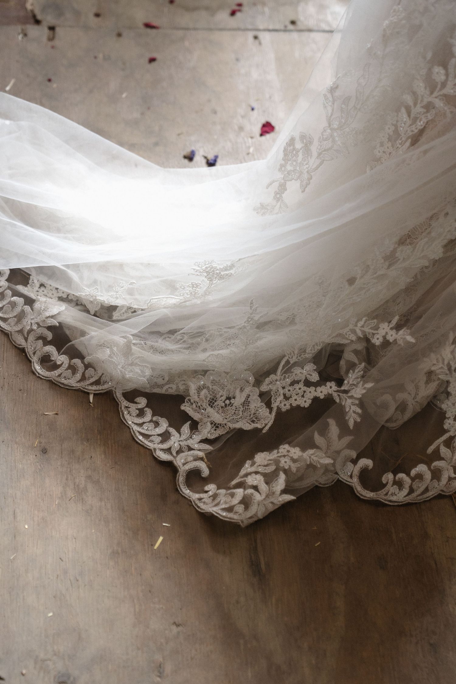 Elegant white lace wedding veil with floral embroidery trailing on a rustic wooden floor with scattered flower petals.