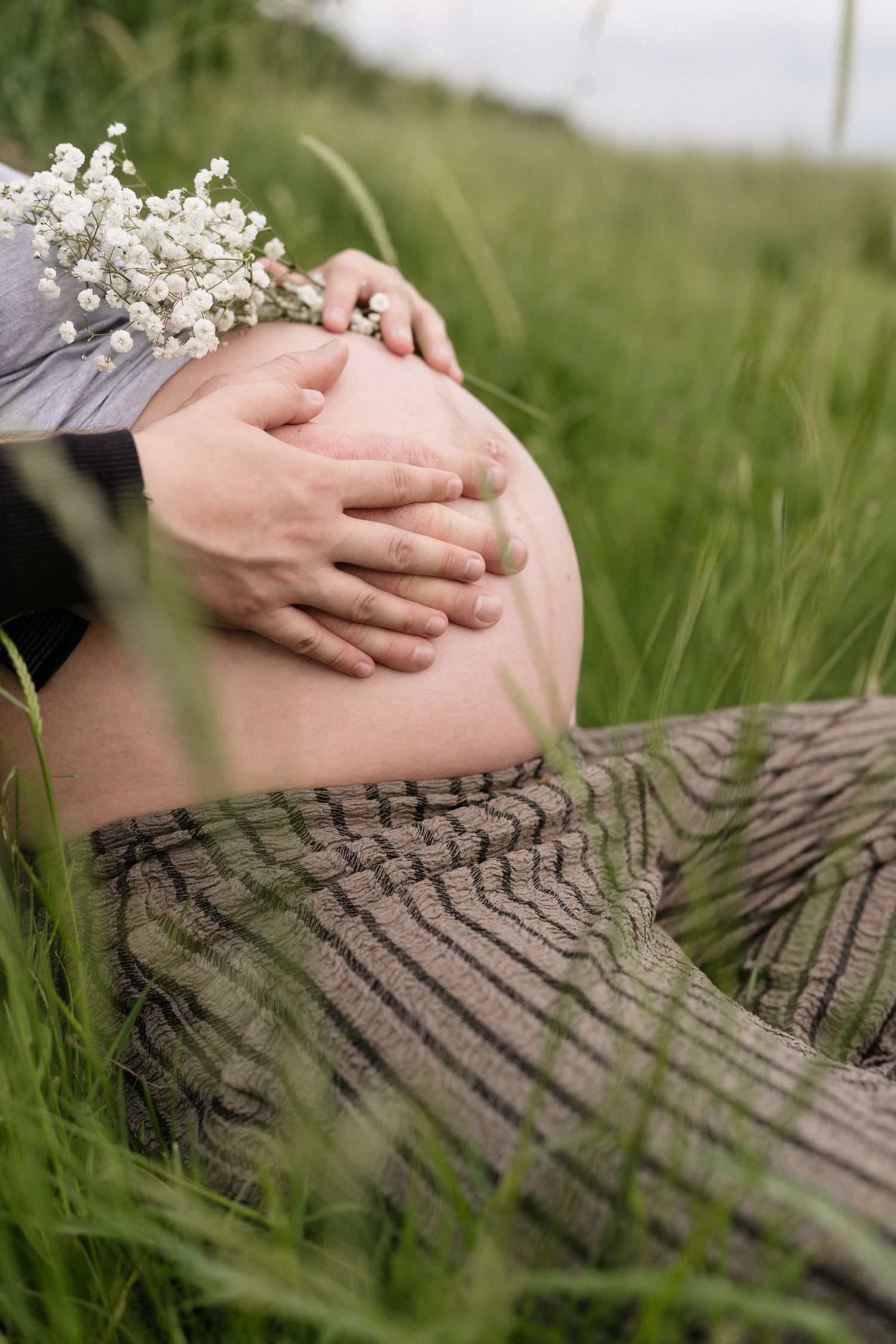 Pregnant woman sitting in green grass field holding baby bump with partner's hands and white wildflowers.