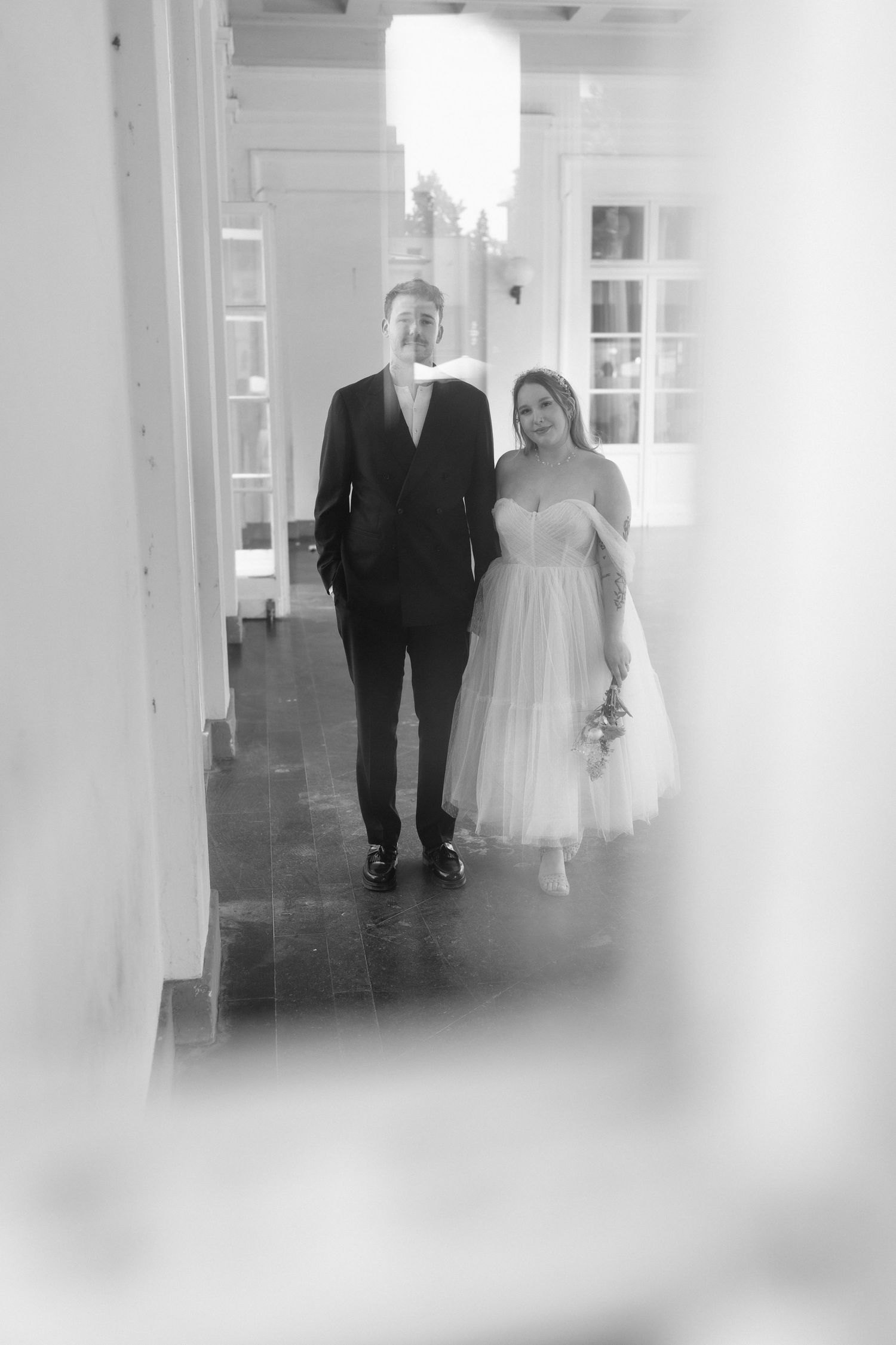 Black and white photo of smiling bride and groom walking together through an elegant indoor corridor.