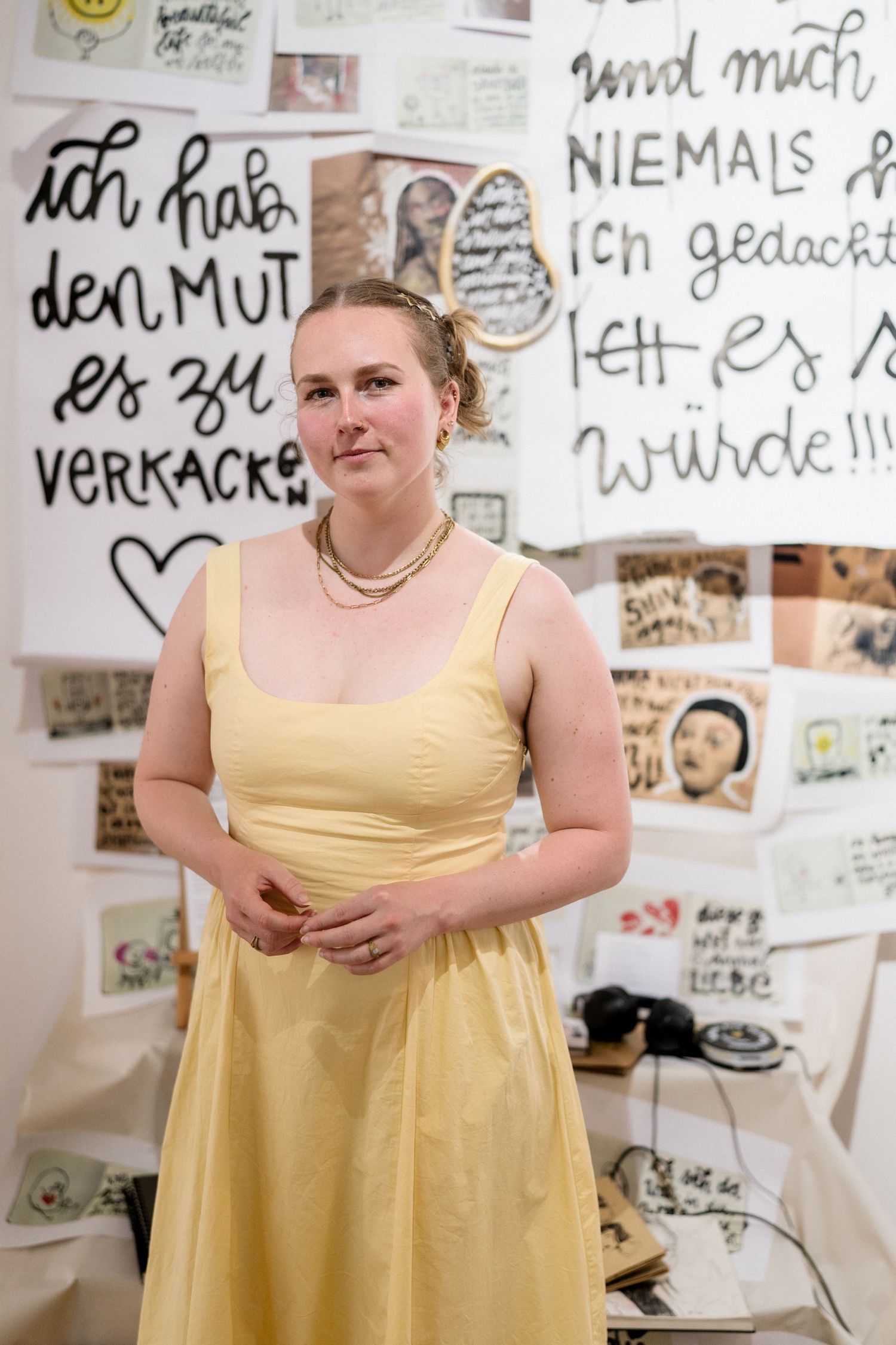 Woman in yellow dress stands before wall covered with German handwritten text and artwork displays.