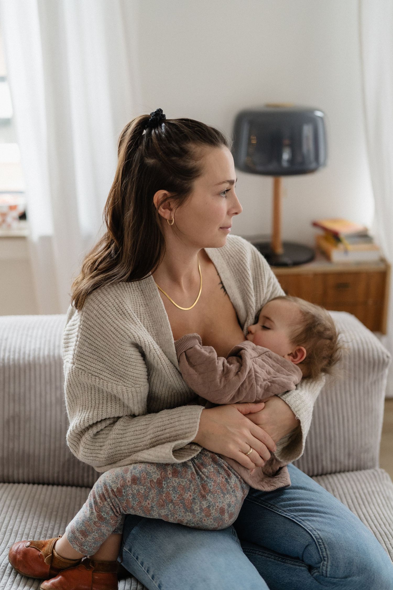 A young mother breastfeeding her toddler while sitting on a couch in a bright, modern living room.