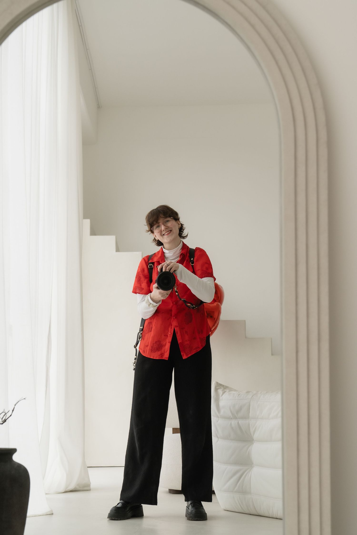 Smiling person in red shirt and black pants holding a camera, standing in a white arched doorway.