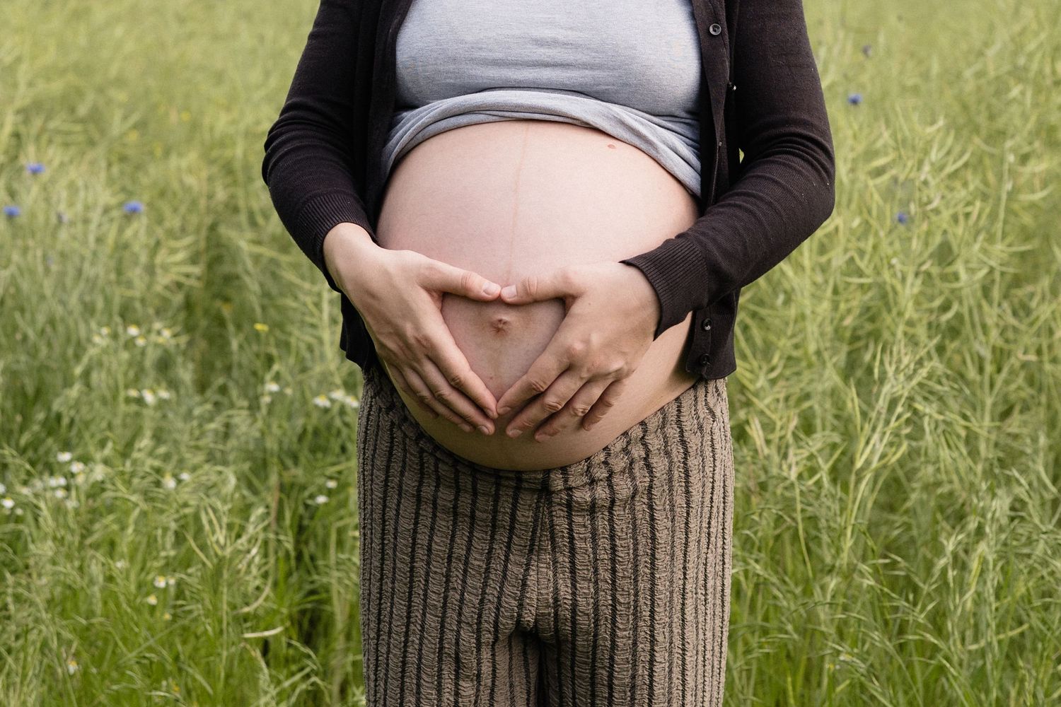 Pregnant woman forming heart shape with hands on bare baby bump in green field.