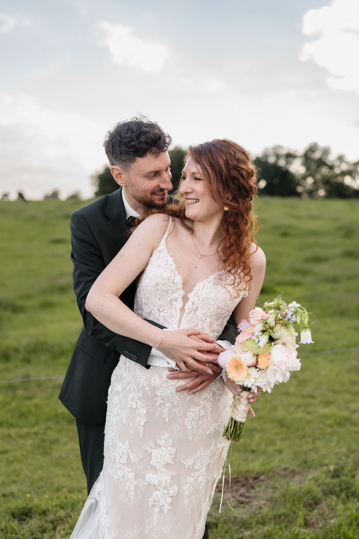 A groom in a black tuxedo embraces his bride in a lace wedding dress holding a pastel bouquet in a green field.
