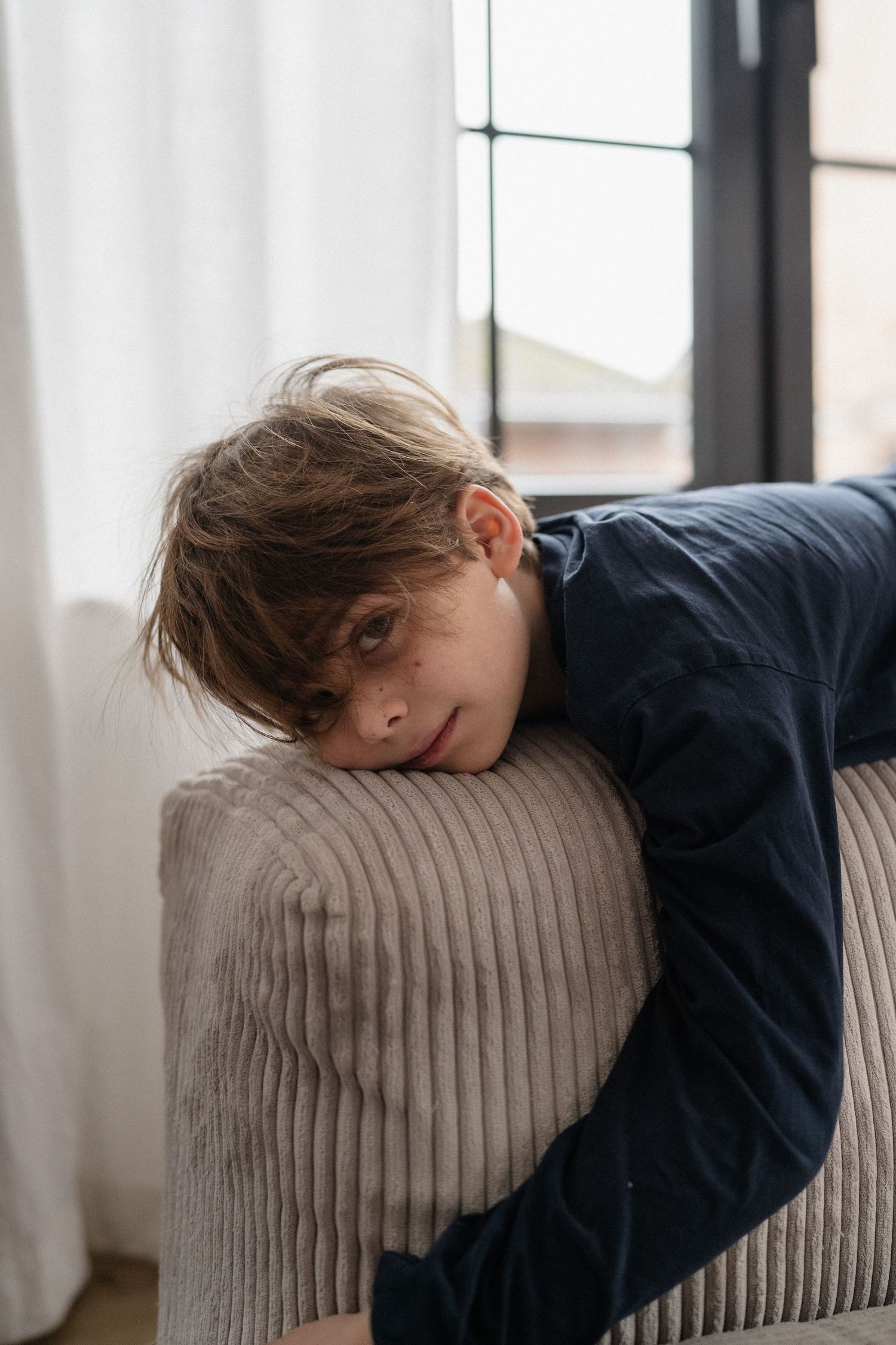 A young boy rests his head on a sofa armrest, looking relaxed near a window with natural light.
