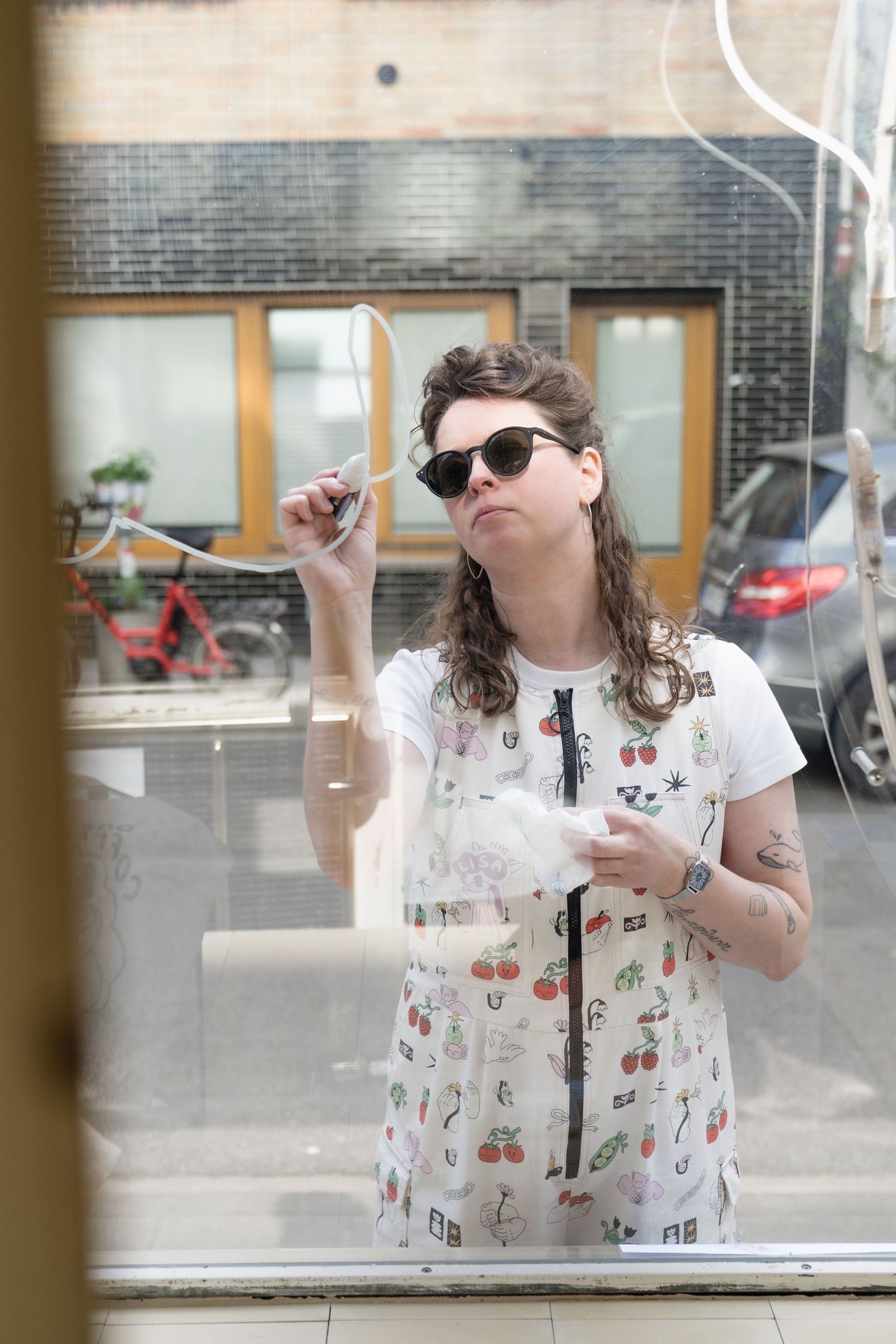 Tattooed woman in patterned overalls cleaning glass storefront window outdoors.