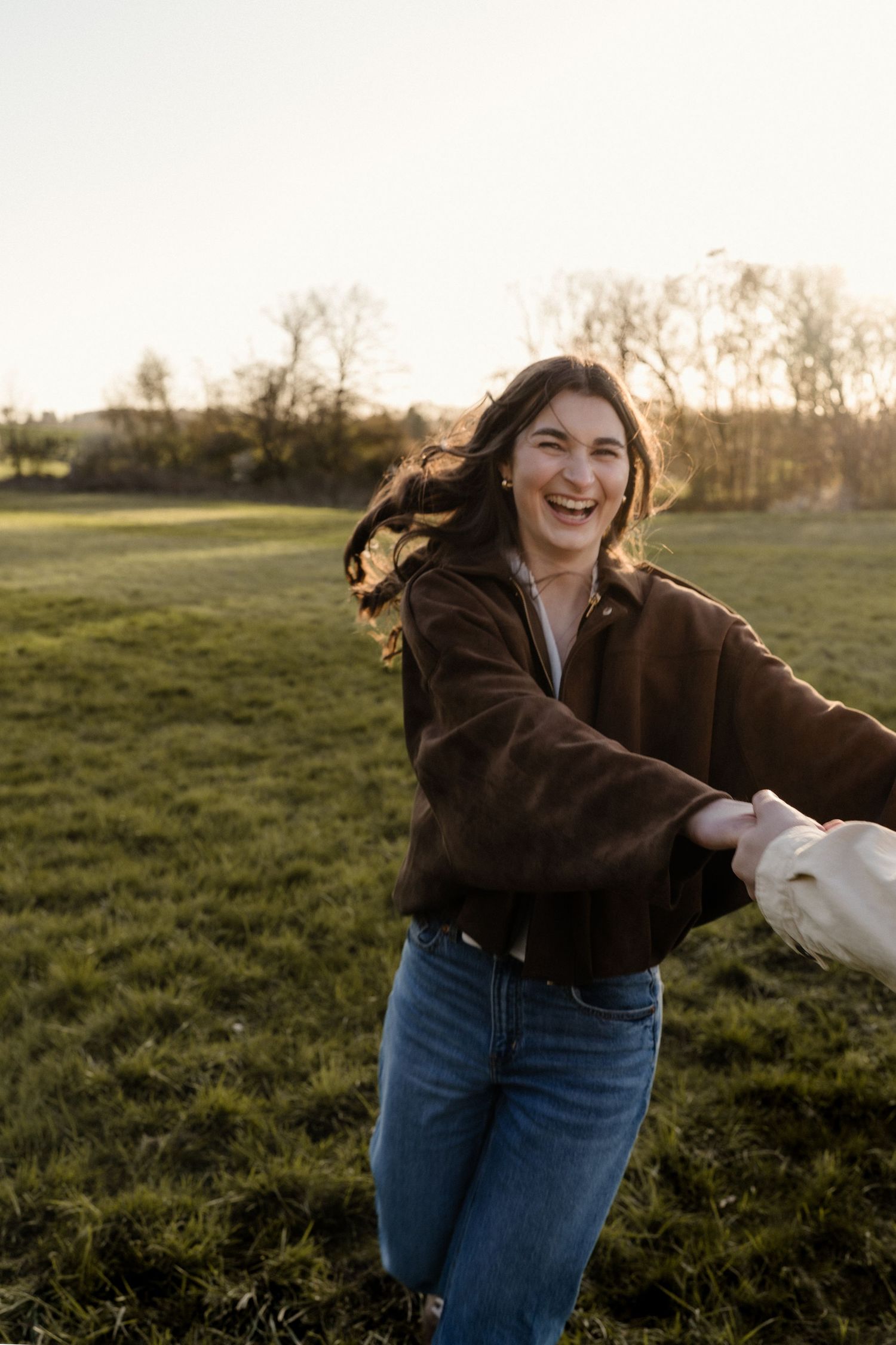 Joyful woman laughing in a green field at sunset, holding hands with partner out of frame.