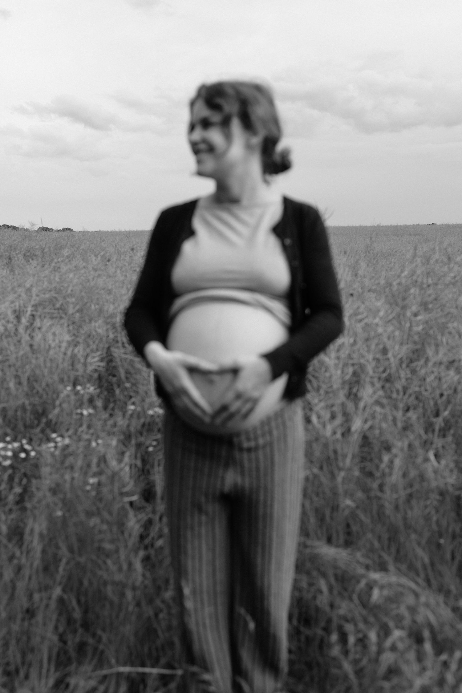 Black and white portrait of smiling pregnant woman making heart shape on baby bump in open field.