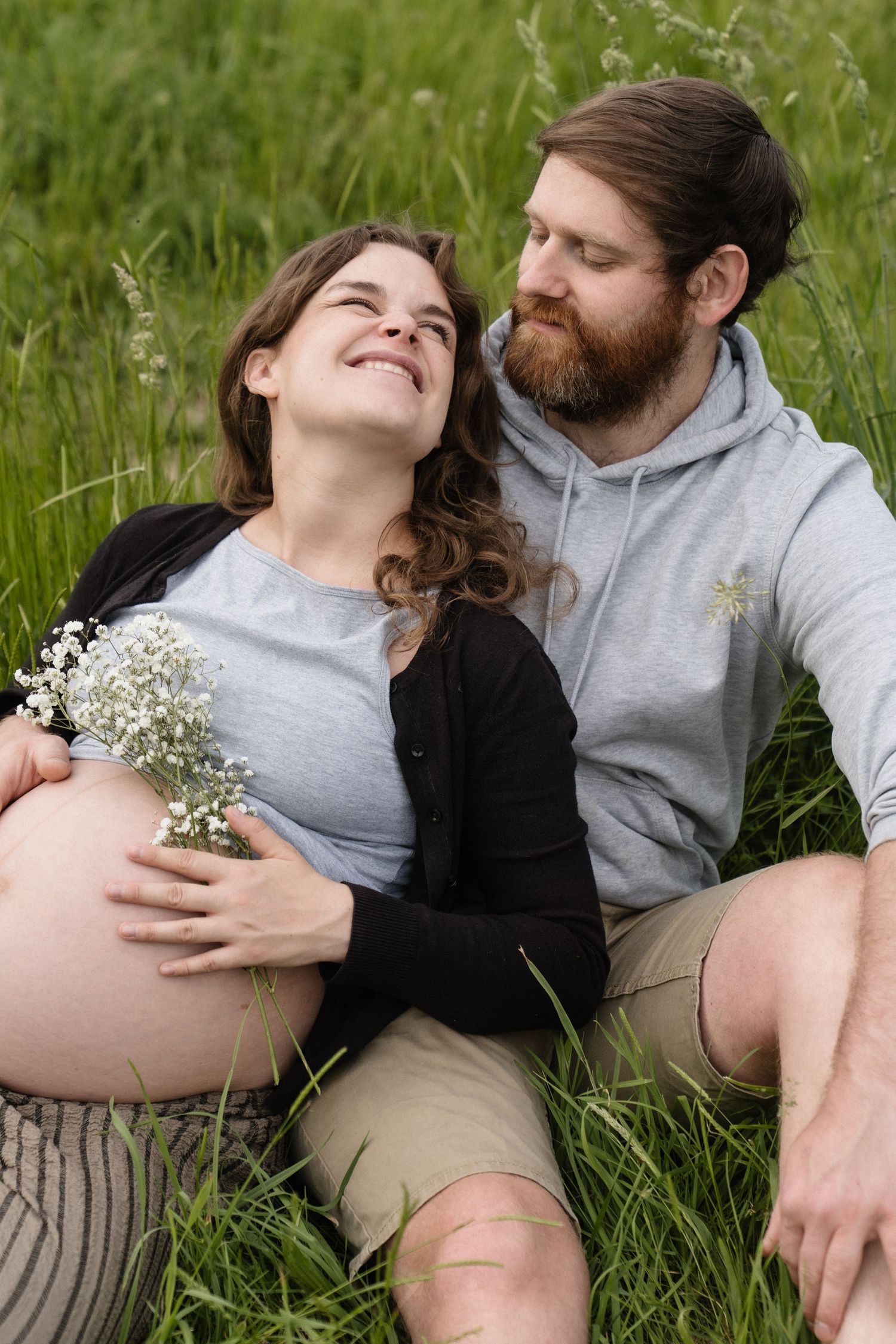 Pregnant woman holding wildflowers sits with bearded man in green grass, both smiling warmly during outdoor maternity photo session.