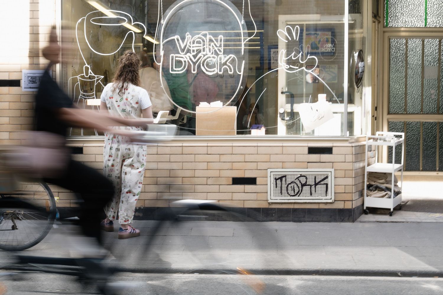 A cyclist blurs past a trendy cafe with Van Dyck neon signage and white tiled counter on a city street.