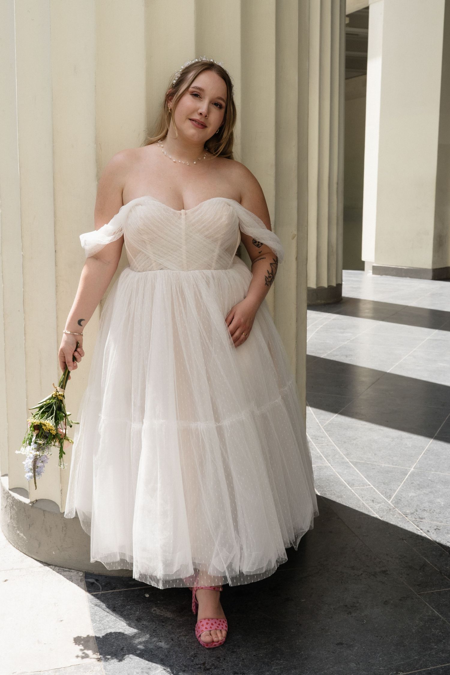 Plus-size bride in off-shoulder blush tulle midi wedding dress holds bouquet between stone columns.