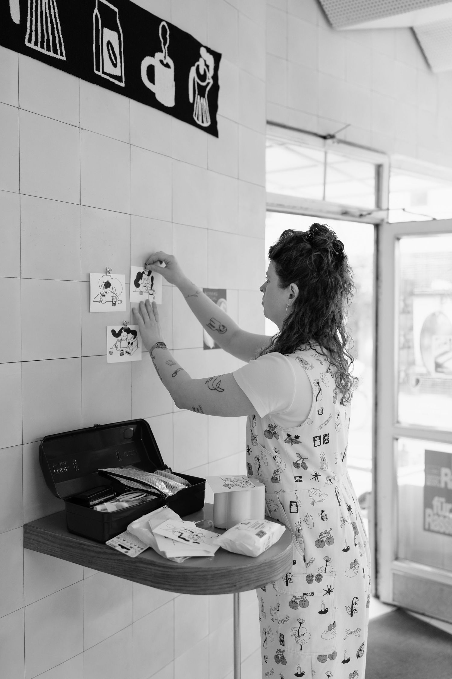 Woman with tattoos arranging art prints on white tiled wall in a cafe interior.