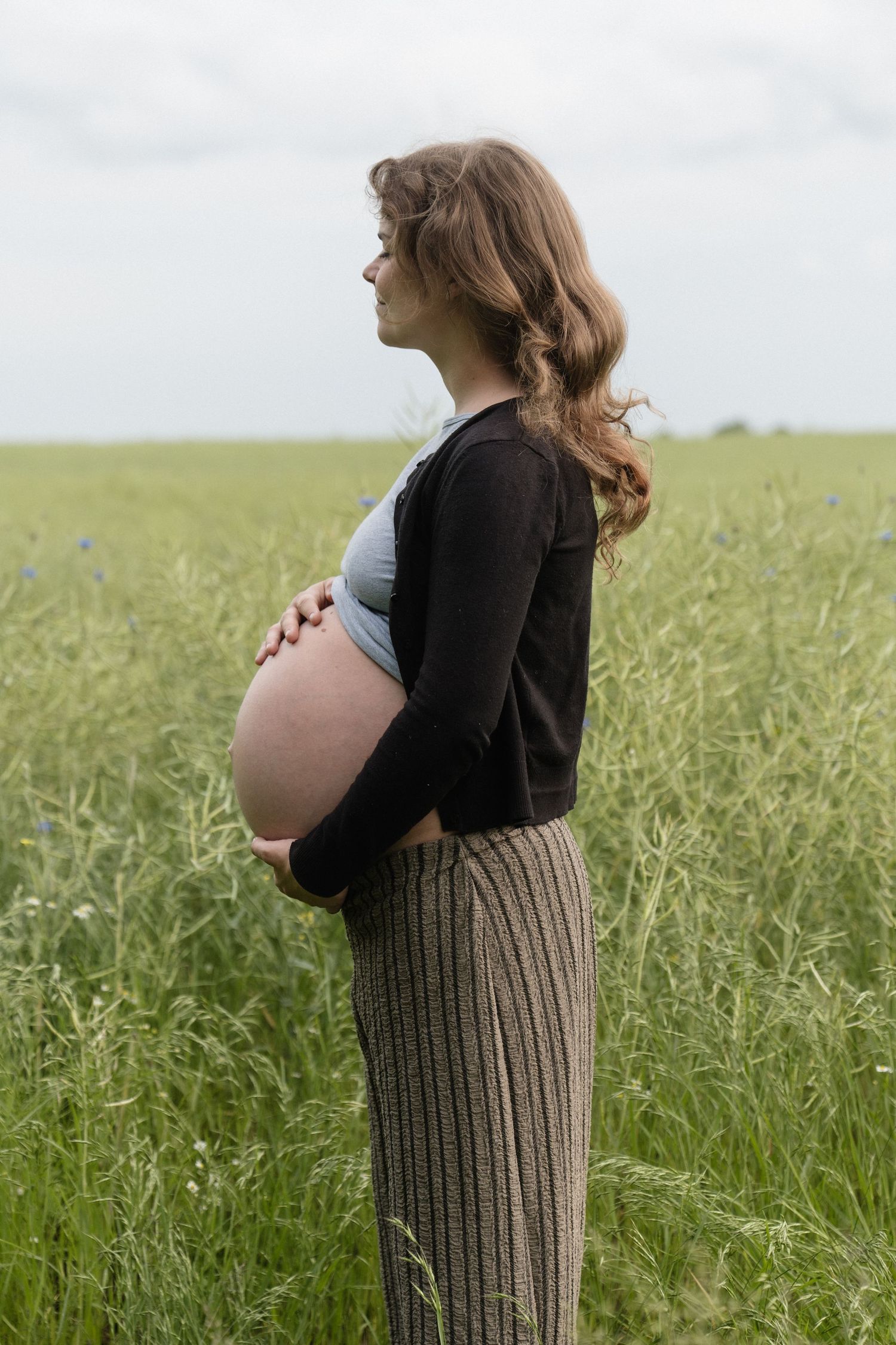 Side profile of pregnant woman cradling baby bump while standing in lush green field.