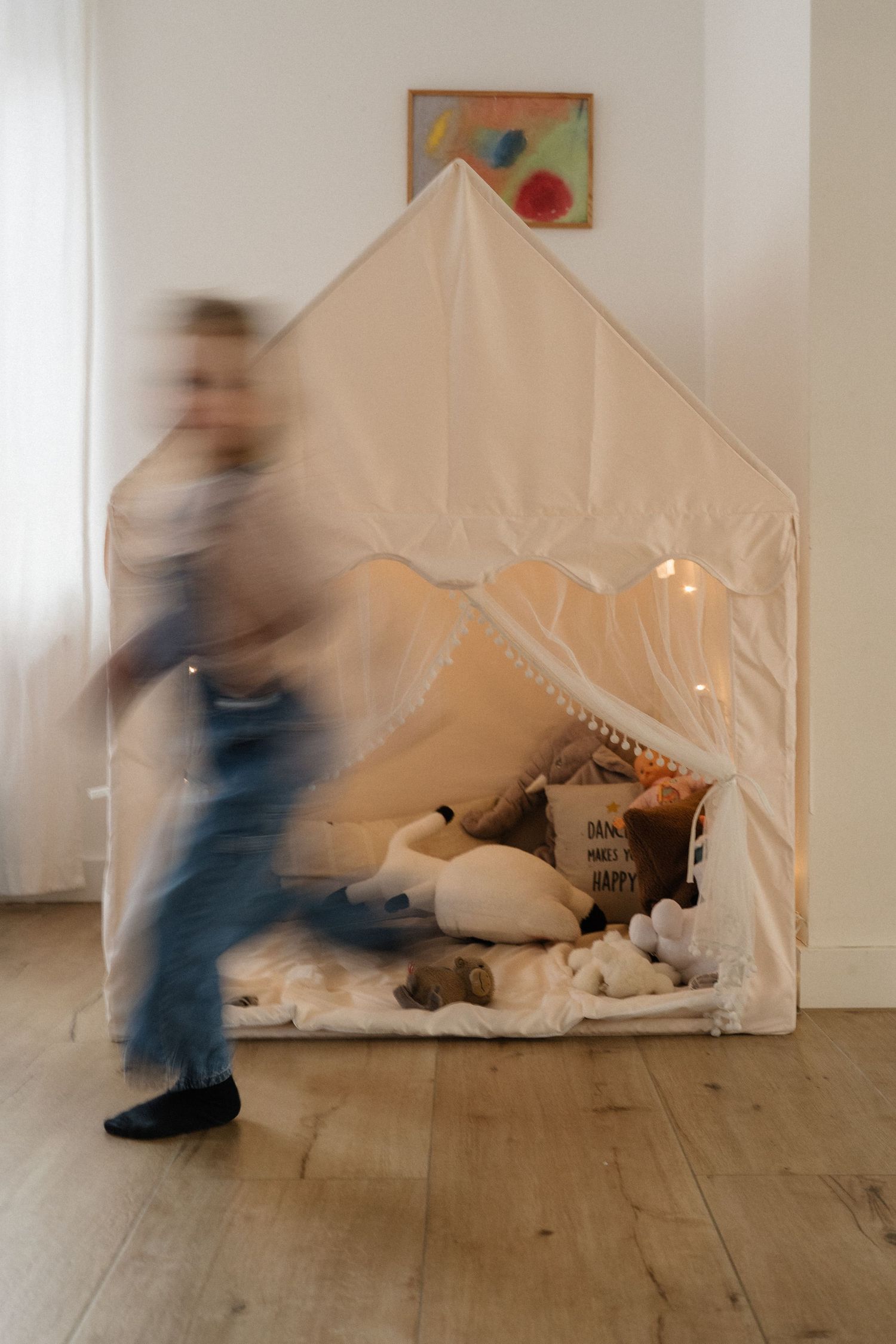 A blurred child runs past a cozy white play tent with fairy lights and stuffed animals indoors.