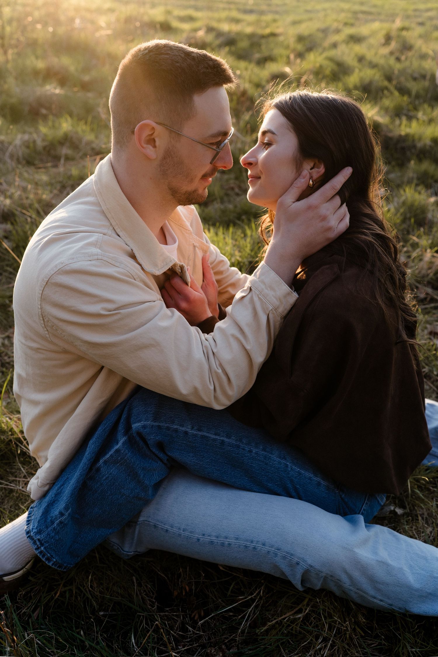 Couple sitting on grass at sunset, smiling and leaning in close, bathed in golden light.