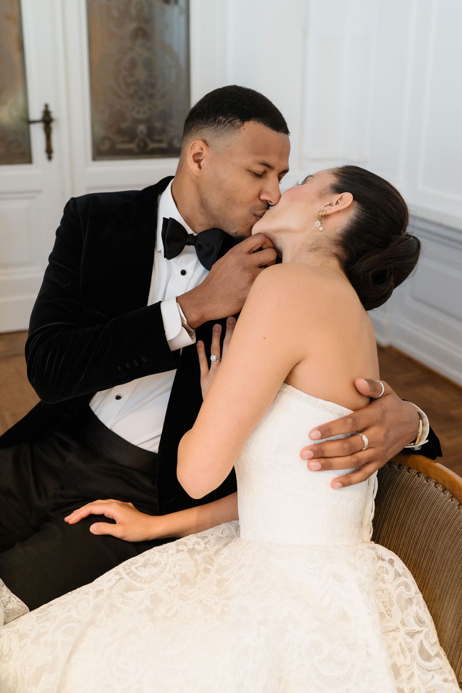 A groom in a black tuxedo tenderly kisses his bride in a white lace wedding gown during their wedding celebration.