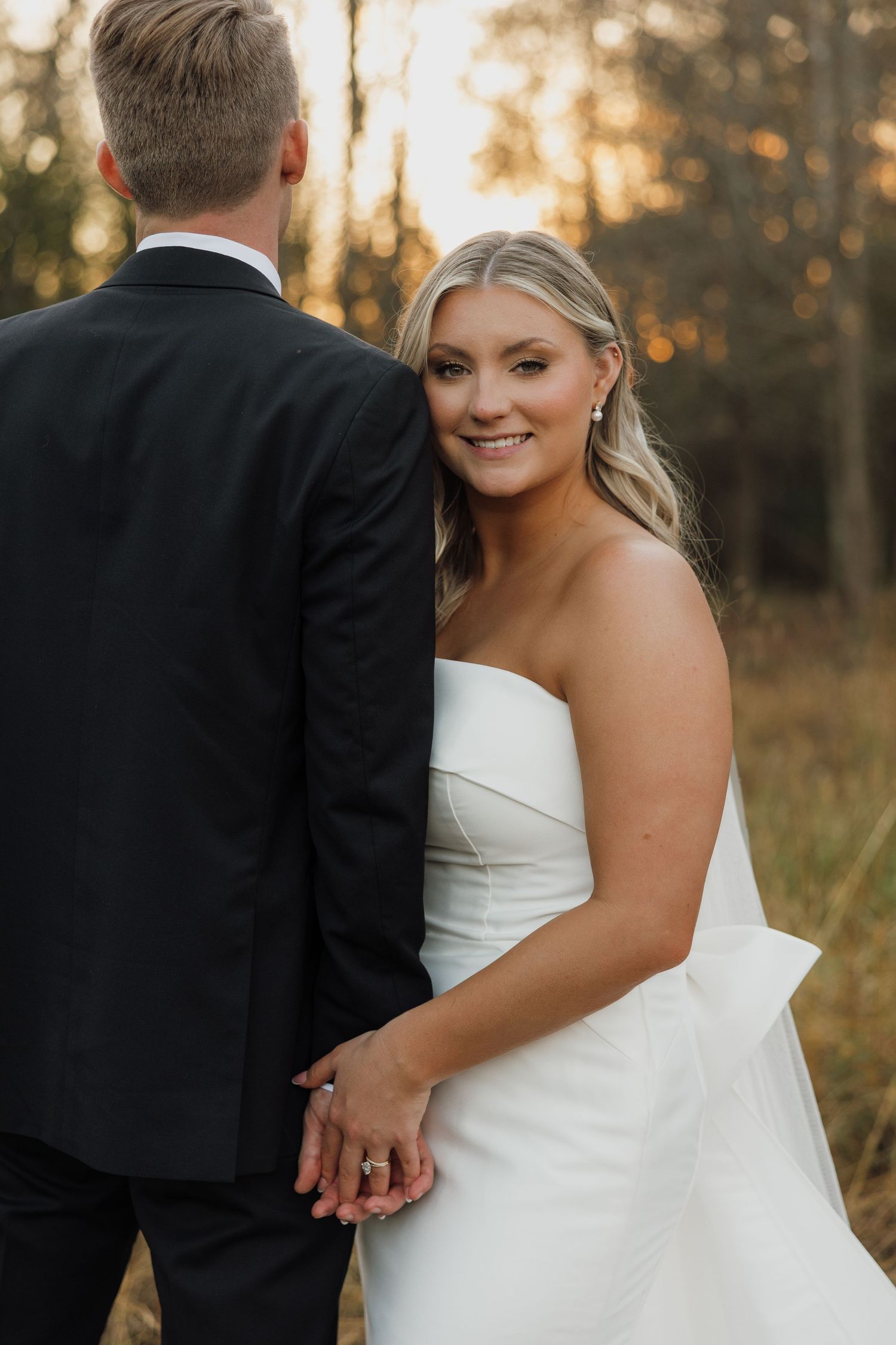 Couple in wedding attire poses together at sunset in a natural outdoor setting with golden light filtering through trees.