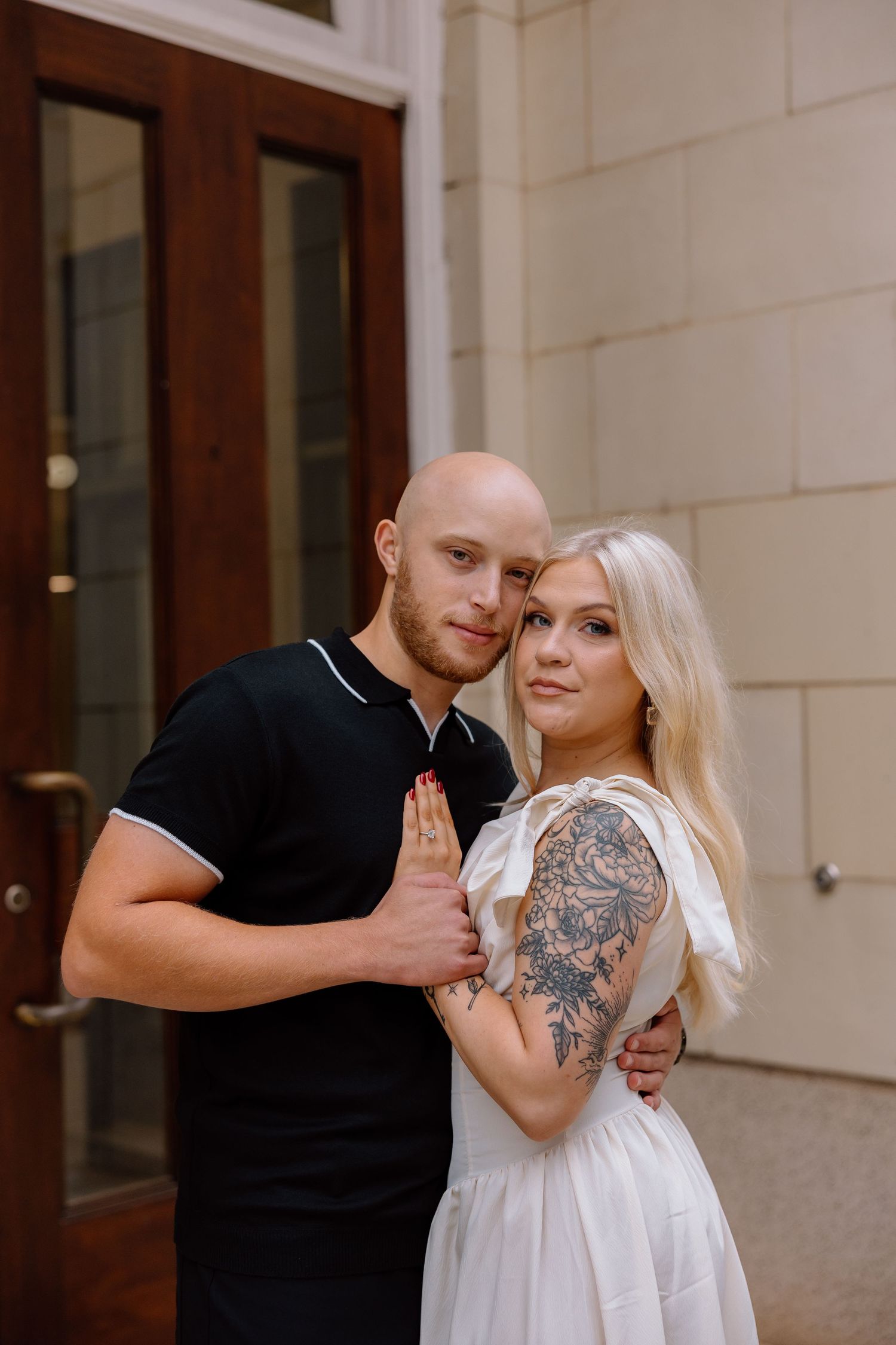 A couple poses together outside a building, with the woman wearing a white dress showing arm tattoos.
