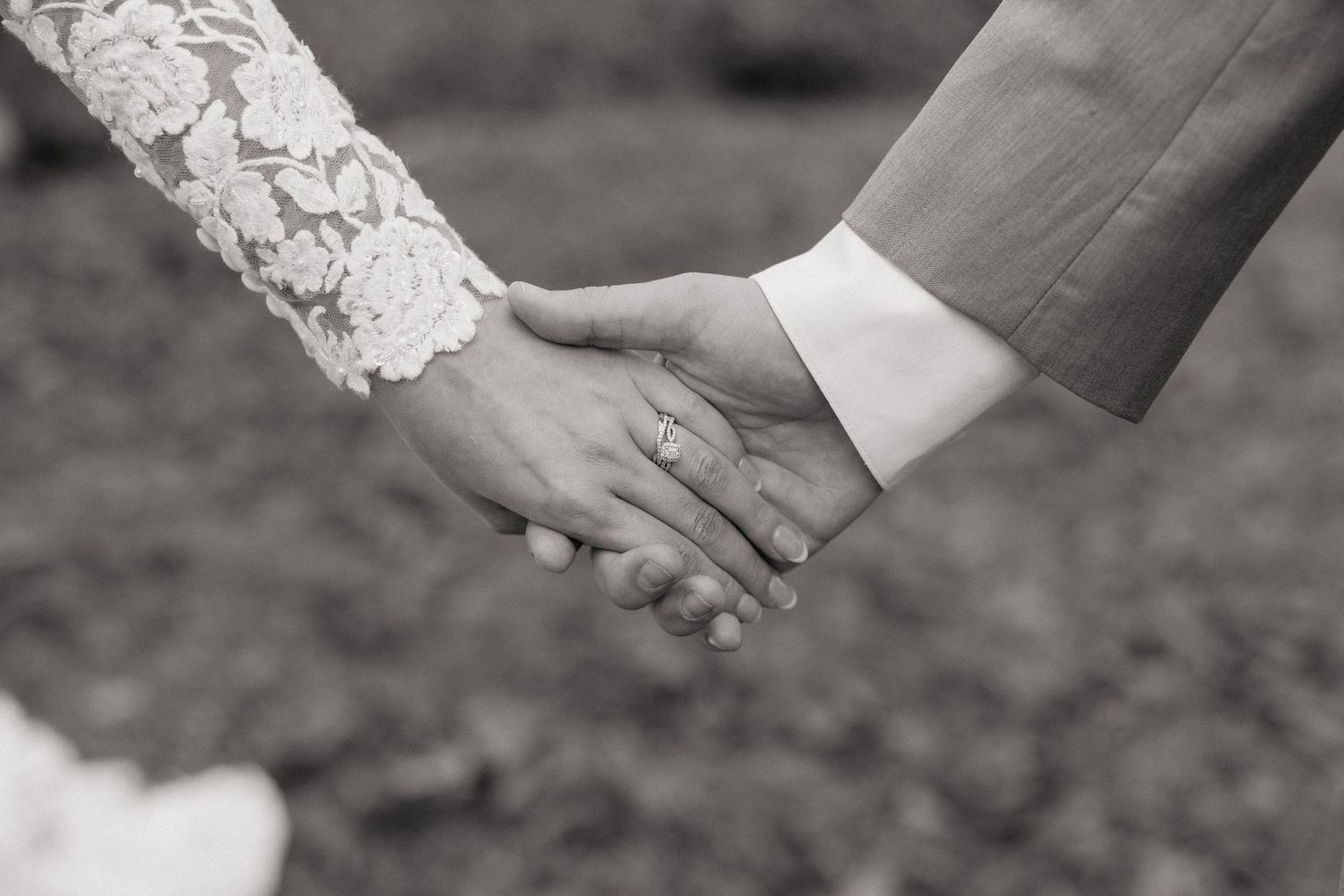 Close-up black and white photo of newlyweds holding hands showing wedding ring and lace sleeve.